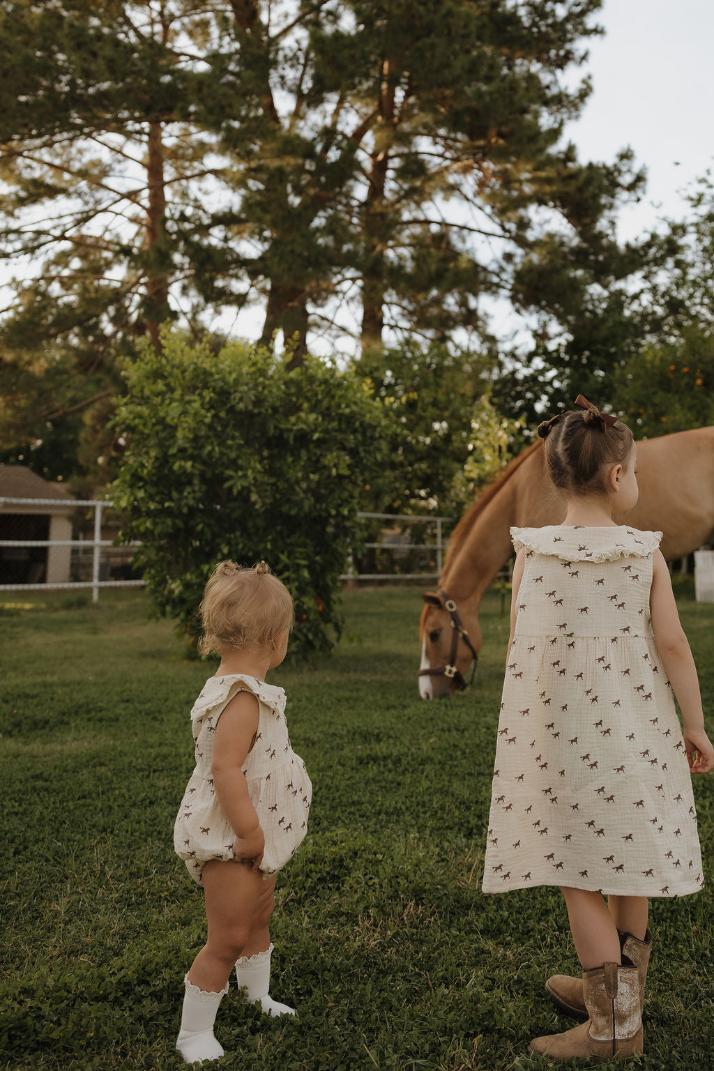 Two young girls wearing forever french baby’s Muslin Romper | Wild Horses stand on grass—one in boots, the other in socks—admiring a brown horse grazing nearby, with tall trees and a fence completing this peaceful scene.