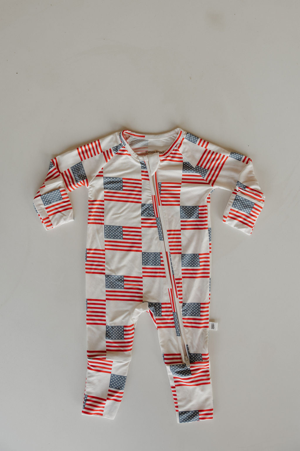 The forever french baby Bamboo Zip Pajamas | Americana feature long sleeves and legs, an all-over American flag pattern, and a diagonal zipper from neck to left leg, shown laid flat on a light background.