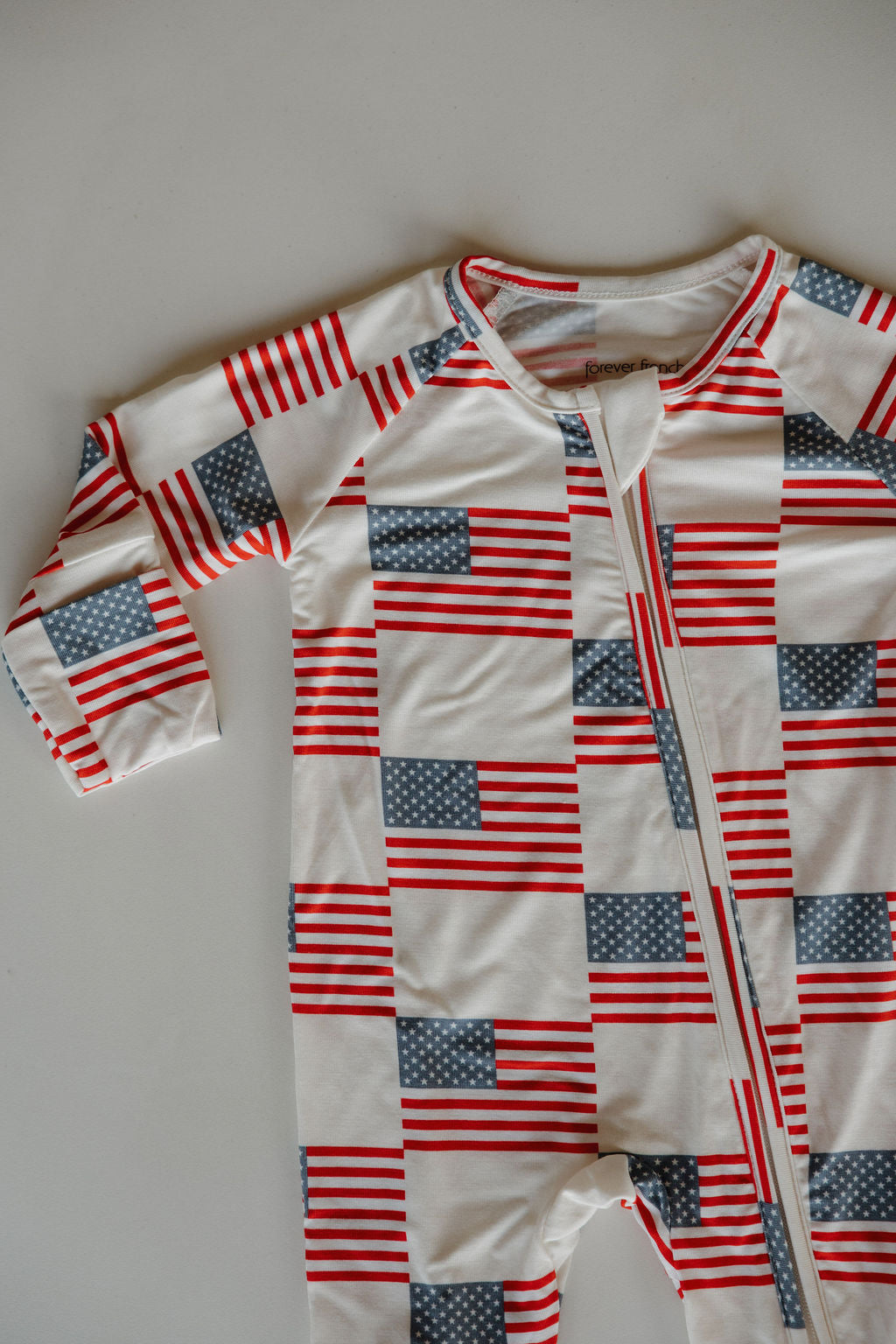 The forever french baby Bamboo Zip Pajamas | Americana is a long-sleeve onesie featuring an American flag pattern on a light background. Made from soft, hypo-allergenic bamboo, it includes a diagonal zipper from the neckline to the left leg.