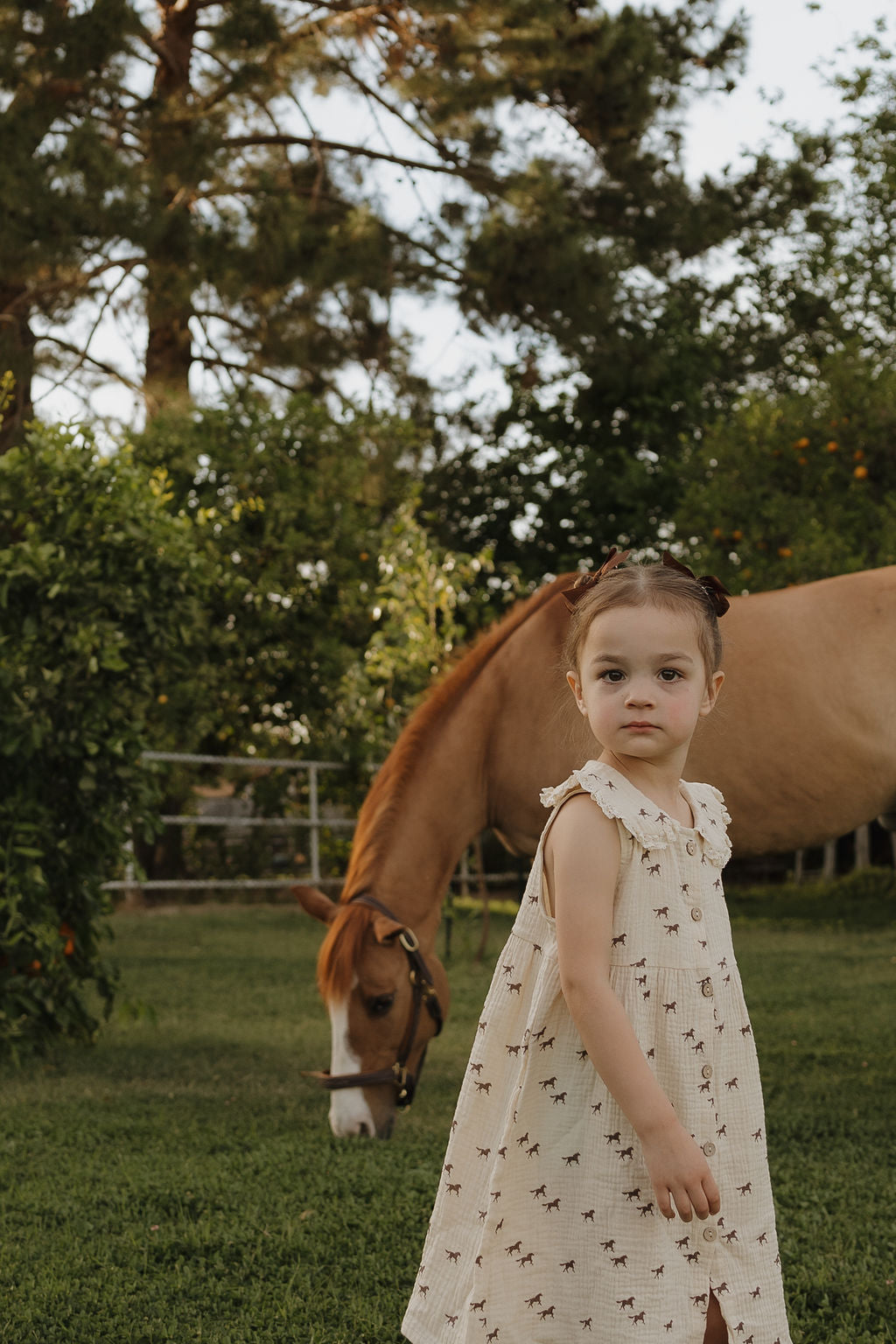 A girl wears the forever french baby "Child Muslin Dress | Wild Horses," standing on grass with a brown horse grazing behind her among lush trees on a sunny day.