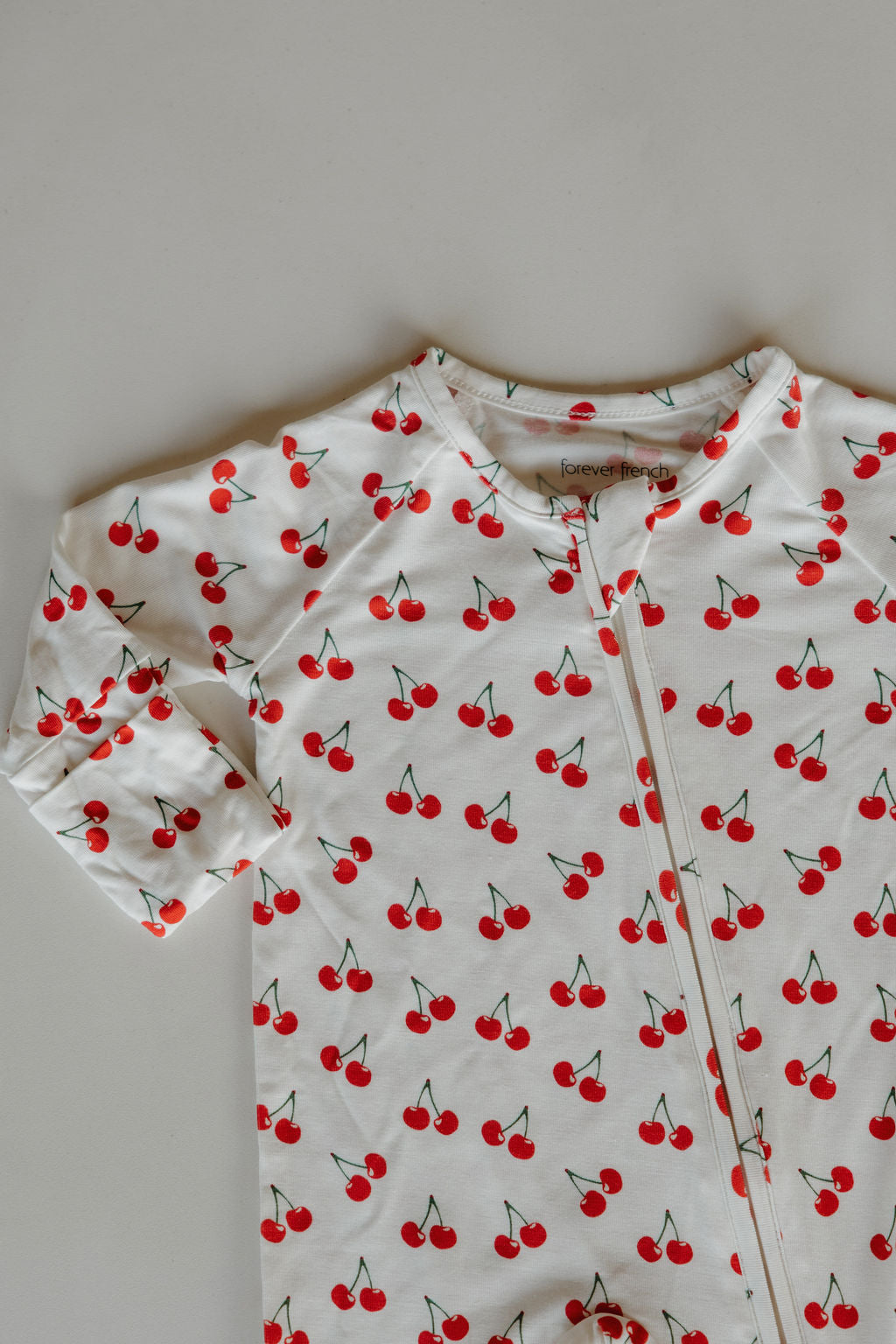 The forever french baby Bamboo Zip Pajamas | Sweetheart Cherries are white, breathable onesies with a zipper, adorned with a scattered red cherry and green stem pattern.