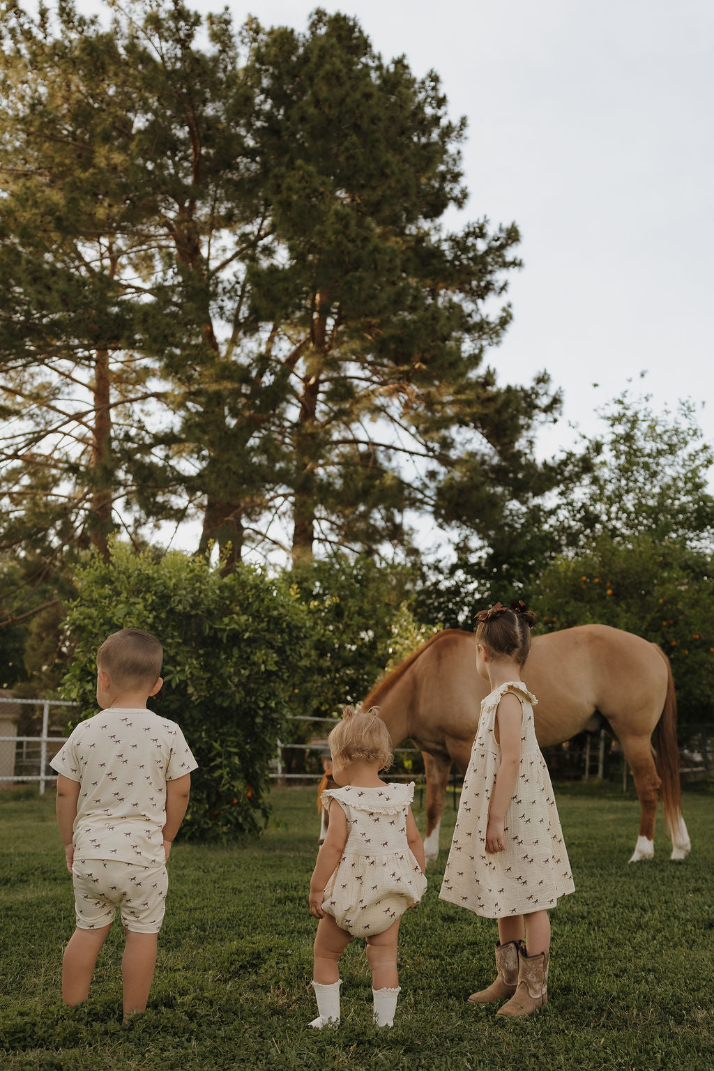 Three young children in light outfits, one in the forever french baby Muslin Romper | Wild Horses, stand on grass watching a brown horse graze near trees and bushes in a sunny outdoor setting.