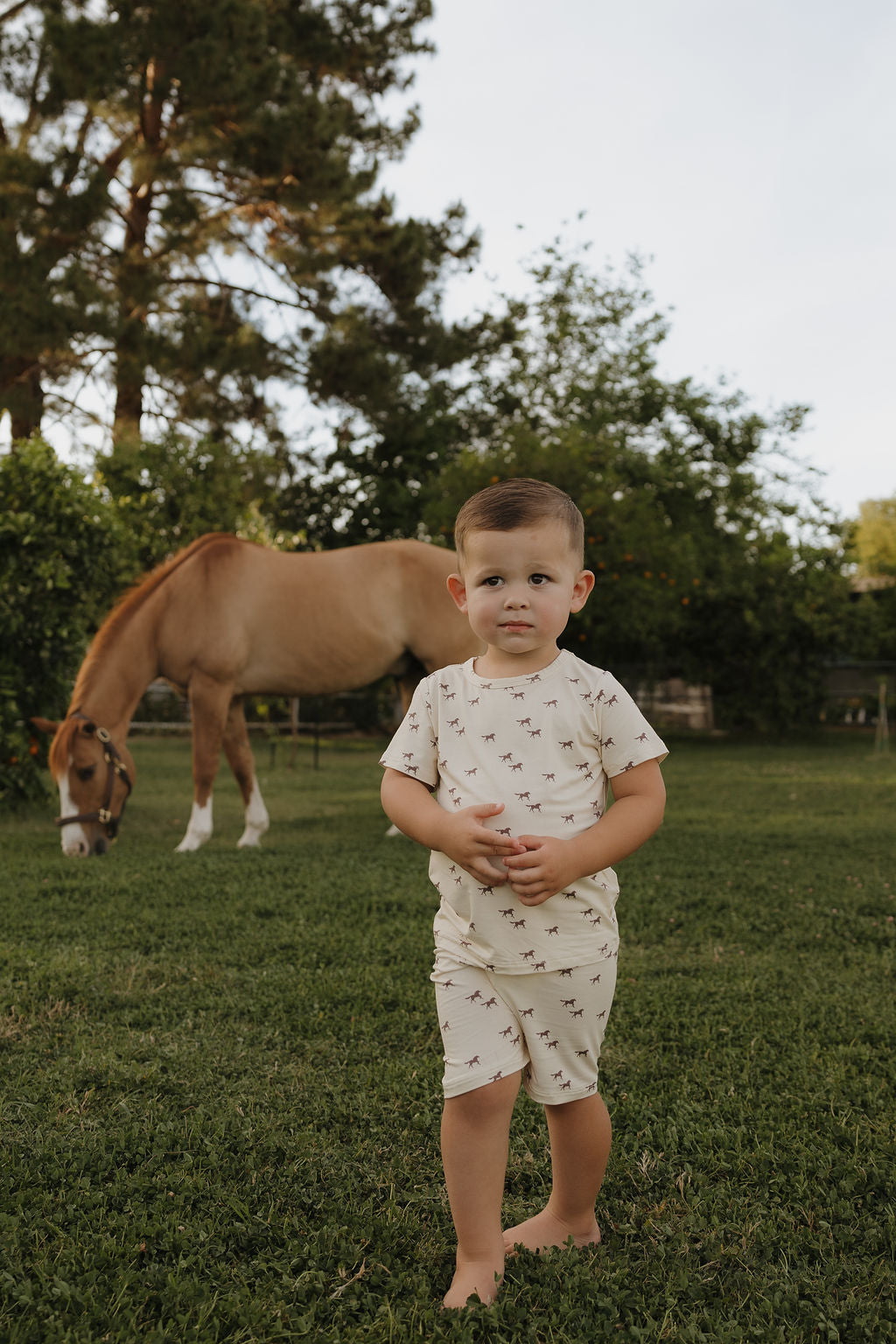 A young child wears the Bamboo Short Set | Wild Horses by forever french baby, standing barefoot on grass near a grazing light brown horse, surrounded by trees and greenery in an outdoor setting.