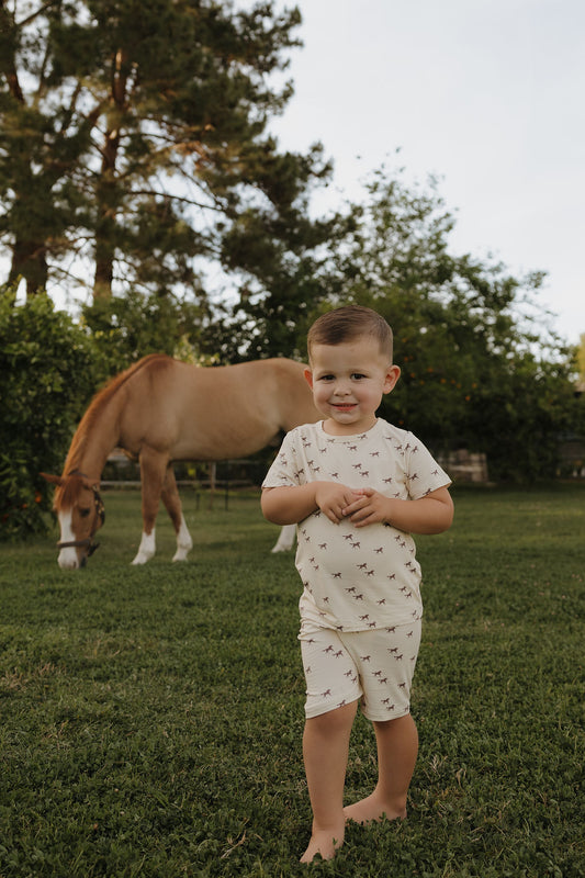 A young child wearing the forever french baby Bamboo Short Set | Wild Horses smiles barefoot on grass, with a brown horse grazing in the background and trees surrounding them.