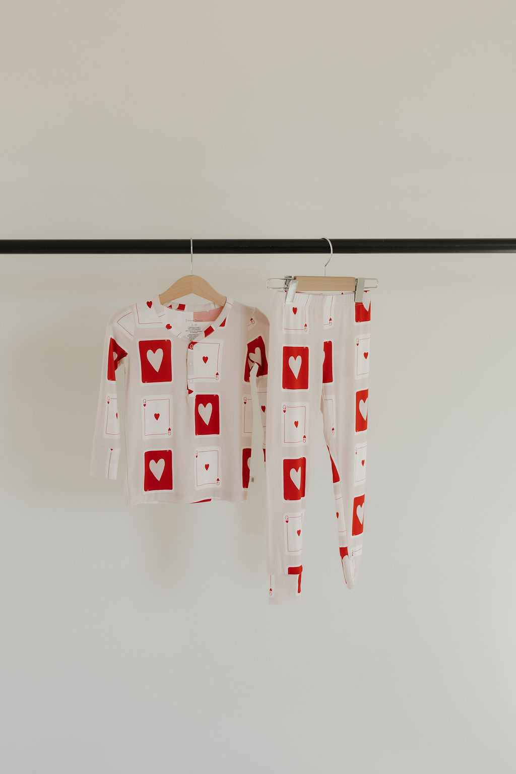 Forever French Baby's "Bamboo Two Piece Pajamas | Love Day" hang on a black clothing rack. These white, breathable pajamas display red hearts and card patterns. The top features long sleeves with buttons, and the pants include an elastic waistband for comfort and style.