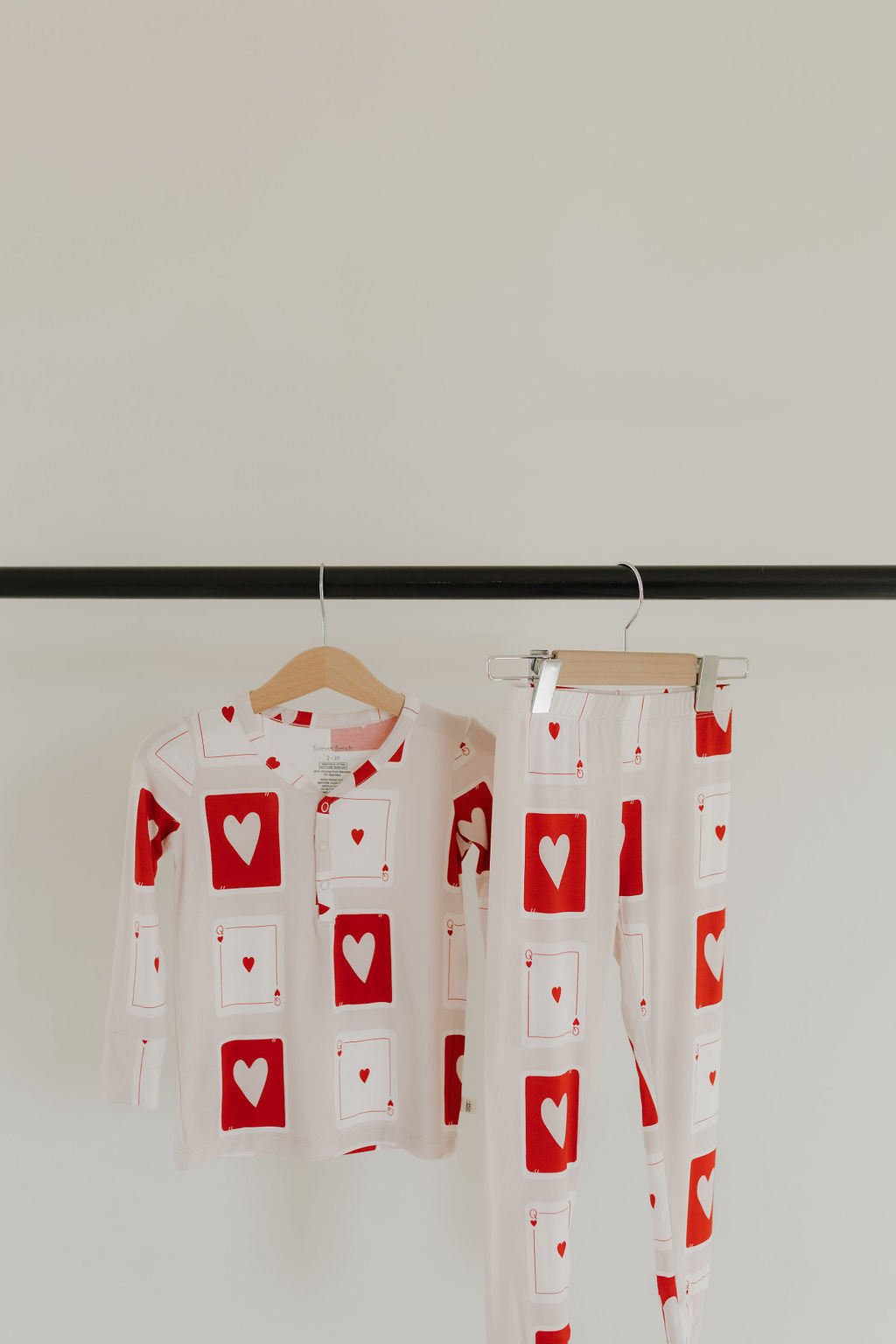 A children's pajama set, the "Bamboo Two Piece Pajamas | Love Day" by forever french baby, hangs on a black rod against a plain background. Made from breathable, hypoallergenic bamboo fabric, it features red hearts and rectangles on white. The set includes a long-sleeve top and pants on wooden hangers.