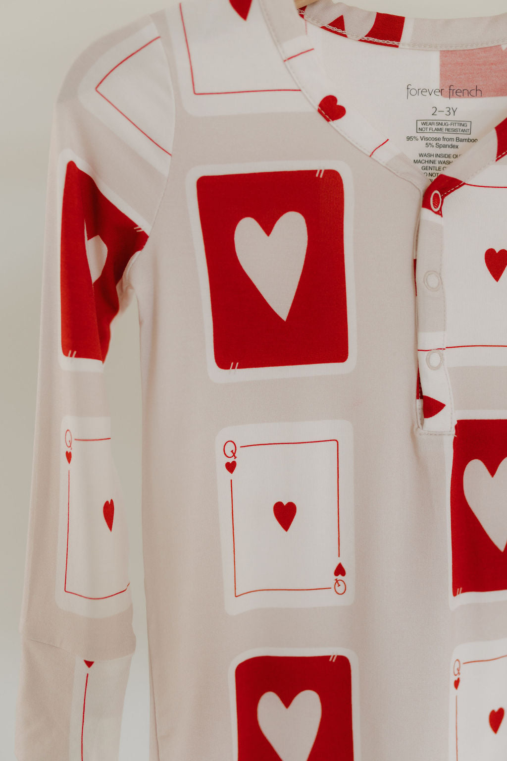 The forever french baby Bamboo Two Piece Pajamas | Love Day features a breathable cream pajama top adorned with red heart designs like Queen of Hearts cards. It has a button-up neckline and long sleeves, perfect for children aged 2-3.