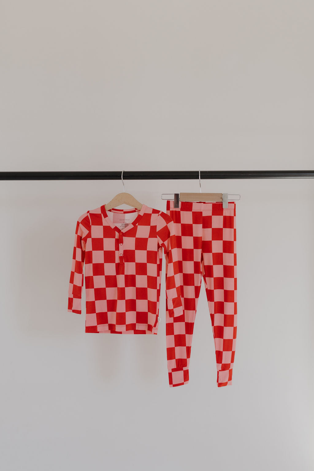 The Bamboo Two Piece Pajamas | XOXO set by forever french baby, featuring a red and white checkered long-sleeve shirt and pants made from breathable bamboo fabric, is displayed on a black rail against a plain background.