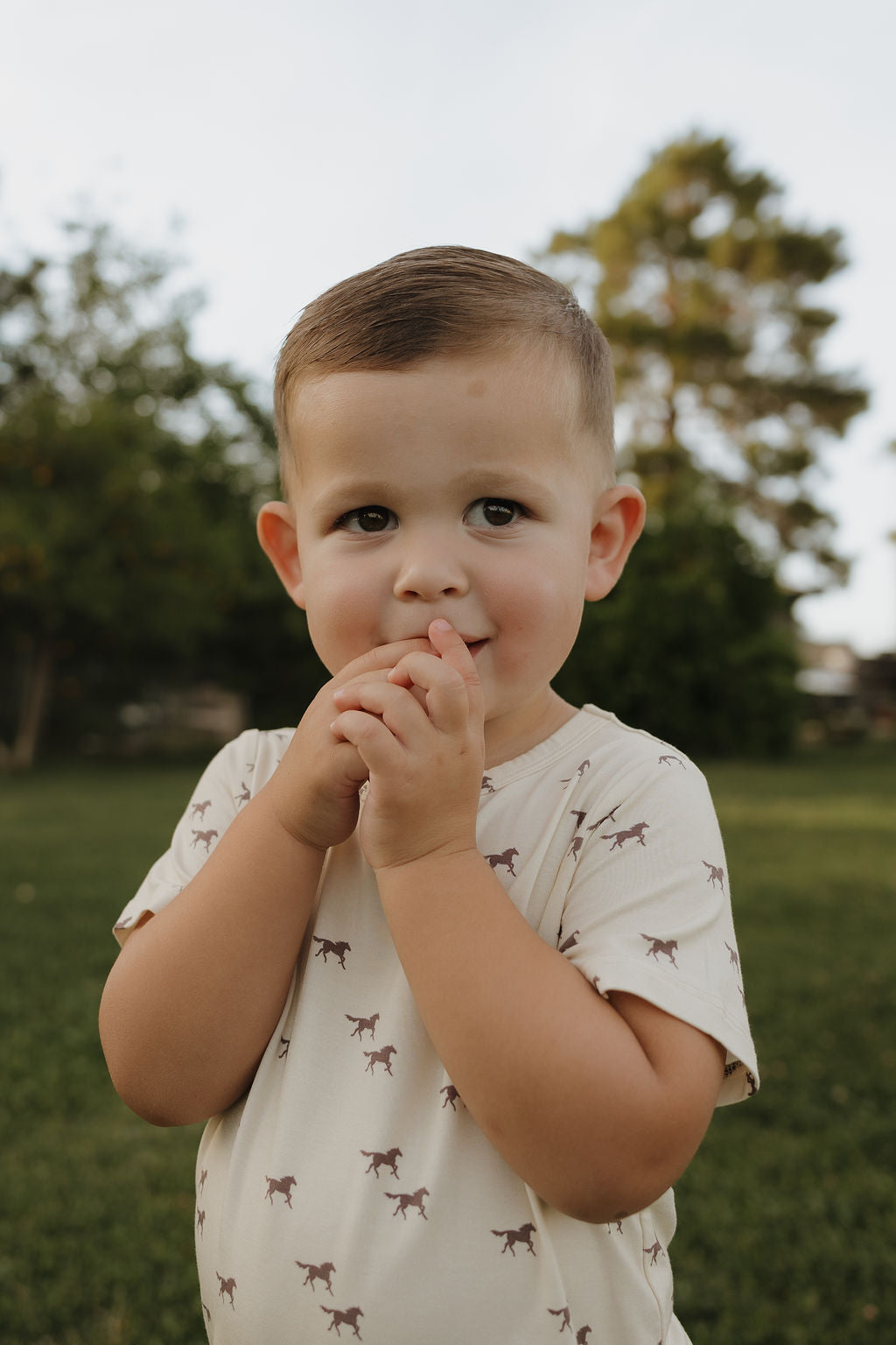A young child smiles softly outdoors on grass, wearing the forever french baby Bamboo Short Set in Wild Horses—a light-colored shirt with animal prints. Trees blur in the background as the child holds their hands near their mouth.
