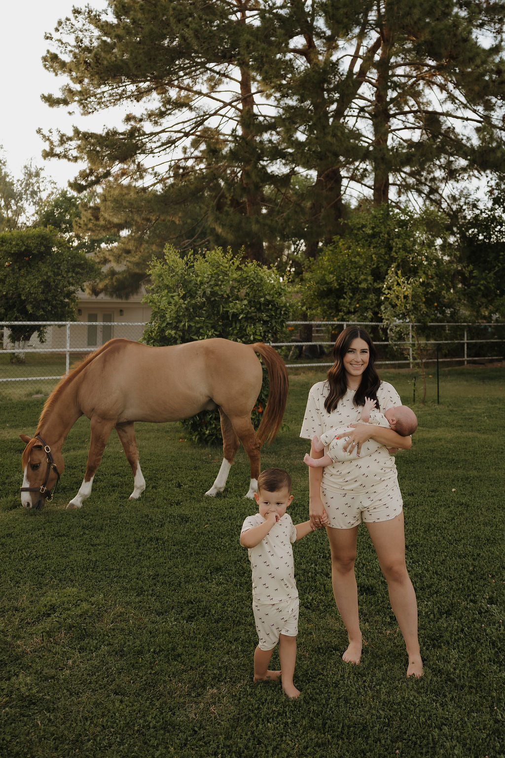 A woman holding a baby stands on grass beside a child in forever french baby's Bamboo Short Set | Wild Horses, with a brown horse grazing nearby. Tall pine trees and a fenced area are visible in the background. Everyone wears light-colored clothing.