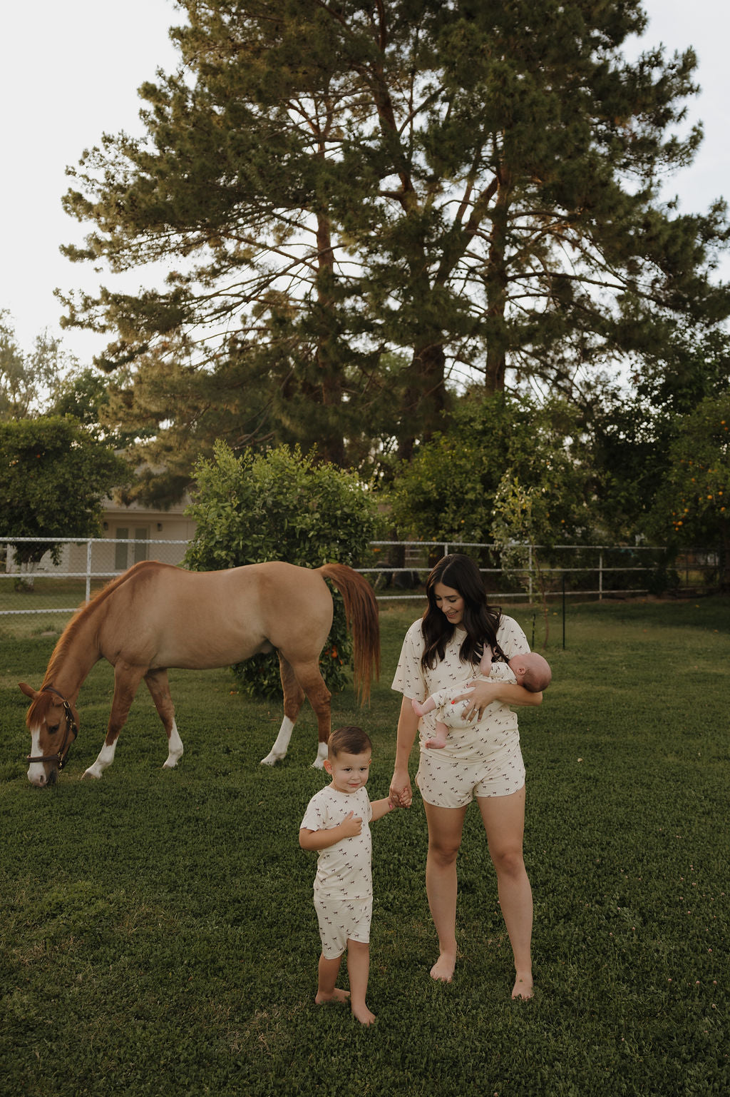 A woman with a baby stands barefoot on grass, holding hands with a young child wearing the forever french baby Bamboo Short Set | Wild Horses. A brown horse grazes nearby; they're outdoors near trees, a fence, and a house.