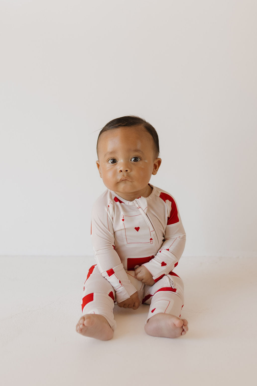 Wearing forever french baby's Bamboo Zip Pajamas | Love Day, a baby poses against a plain backdrop in an adorable white and red one-piece with heart shapes and red patches. Made from breathable fabric, the outfit ensures comfort as the baby gazes at the camera.