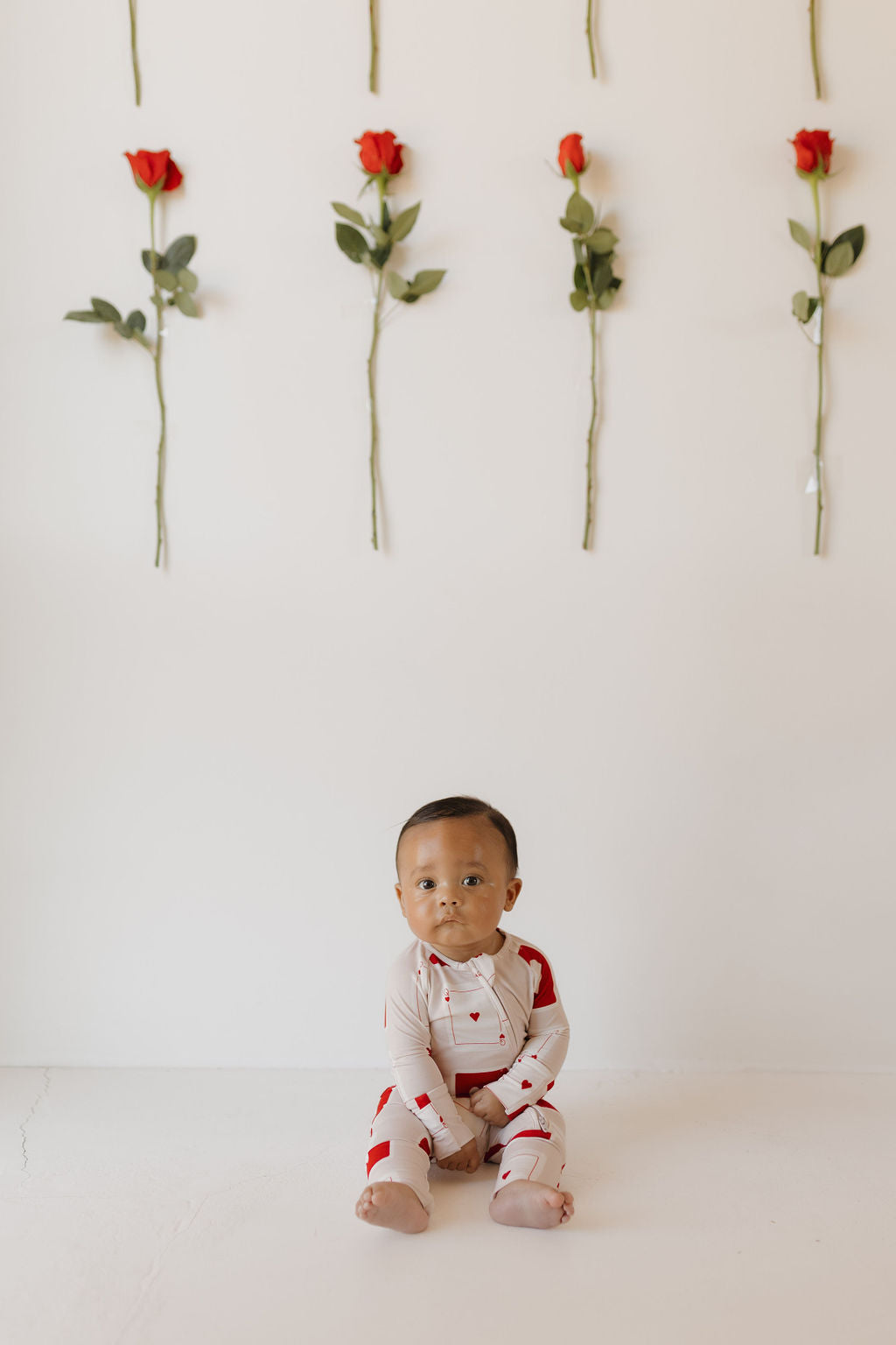 A baby sits on the floor against a white background, dressed in forever french baby's Love Day Bamboo Zip Pajamas. Above, six red roses are vertically arranged on the wall, creating a harmonious and gentle scene.