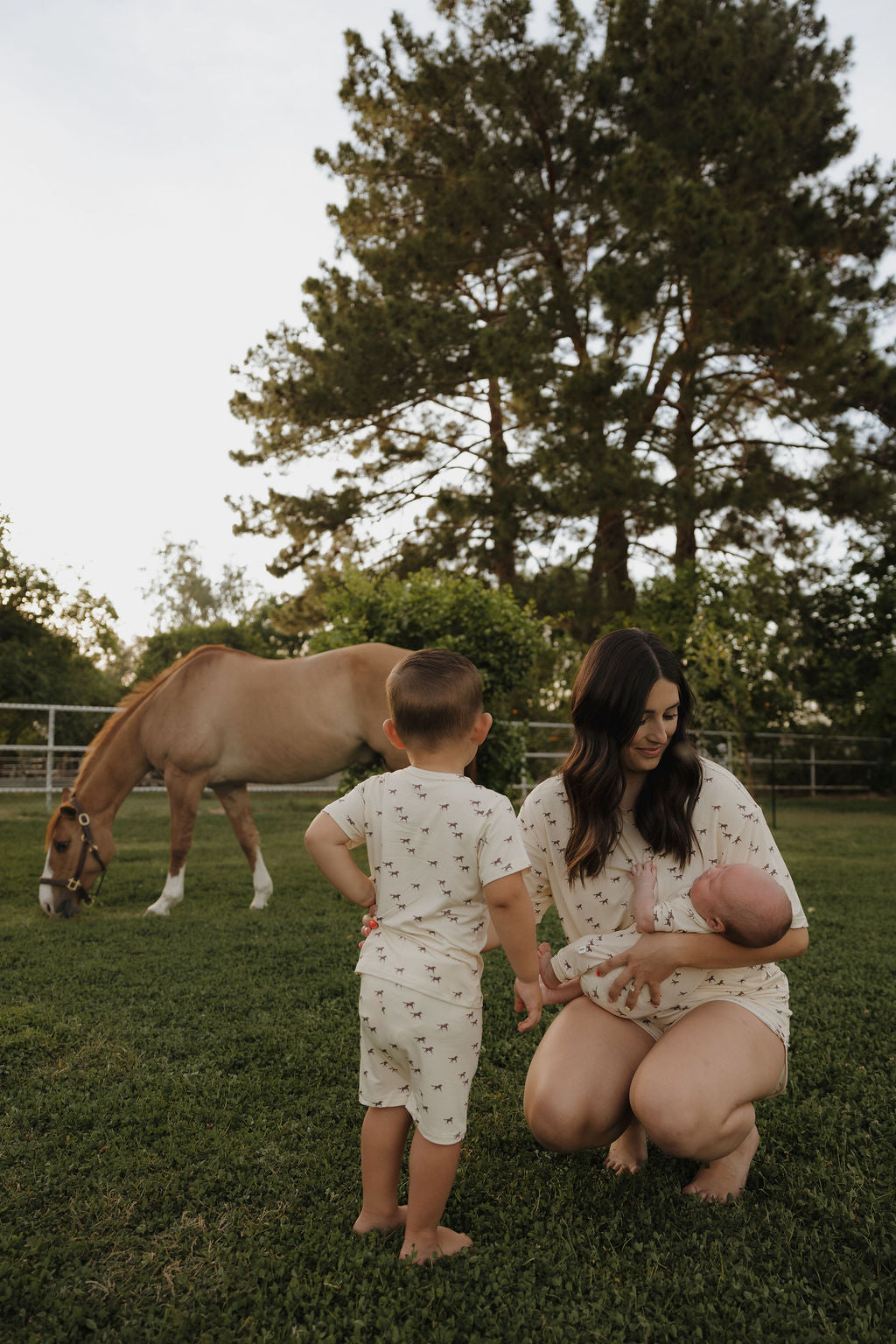 A woman in the forever french baby Women's Bamboo Short Set | Wild Horses kneels on grass holding a baby, as a child stands nearby. A horse grazes and tall trees line the fence in this tranquil outdoor scene.