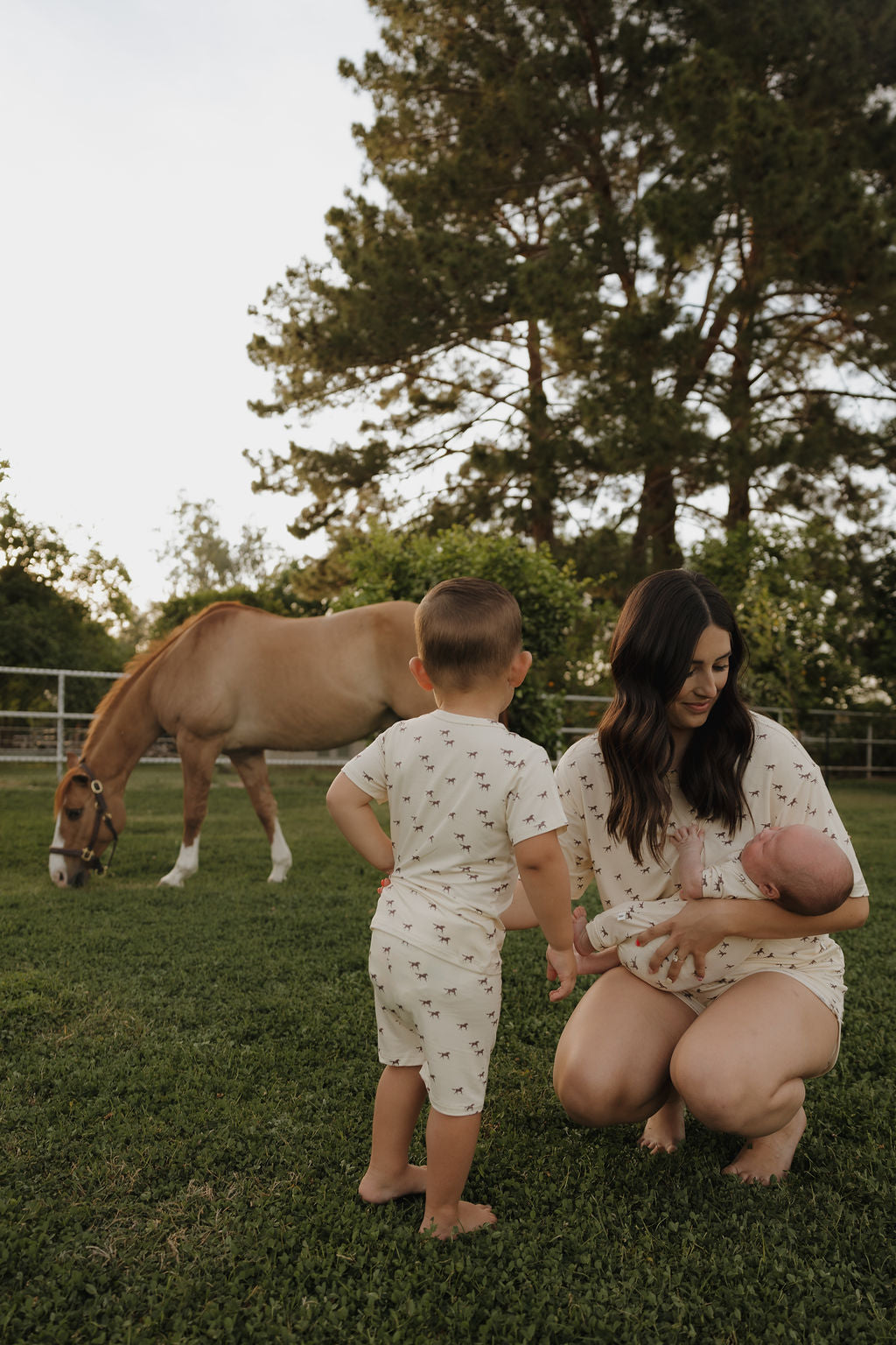 A woman kneels on grass holding a baby while a young child stands beside her. Both children wear the Bamboo Short Set | Wild Horses by forever french baby. A horse grazes nearby among trees and a fence in the background.