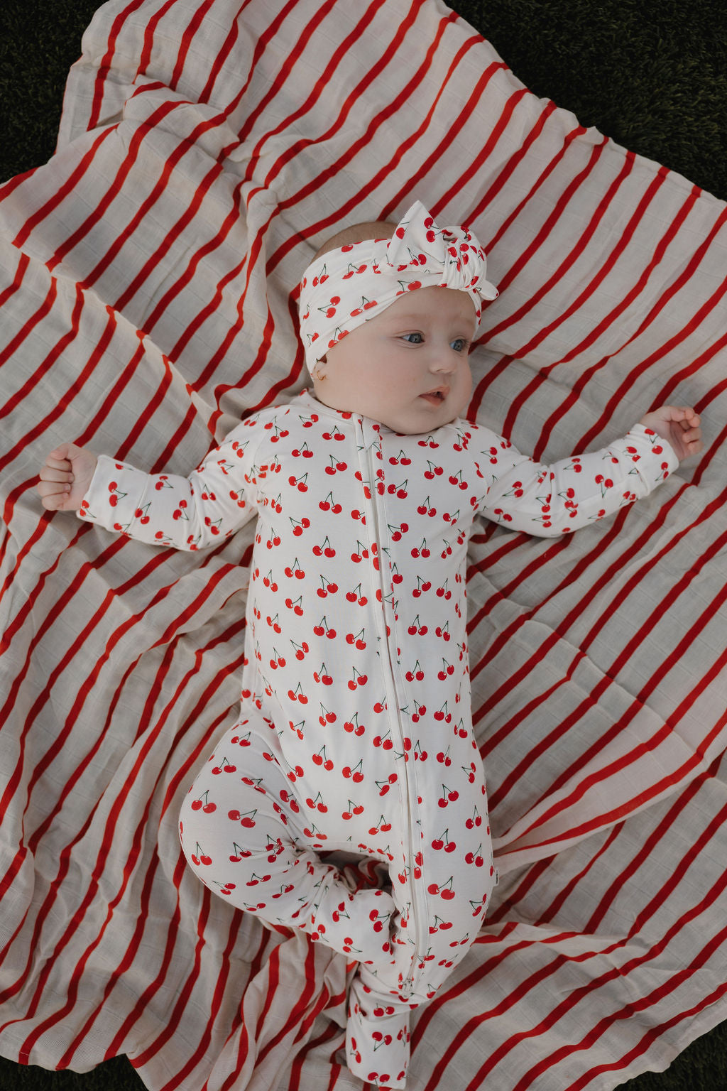 A baby in forever french baby Bamboo Zip Pajamas | Sweetheart Cherries—a white outfit with red cherry prints and matching headband—lies on a white blanket with red stripes outdoors.