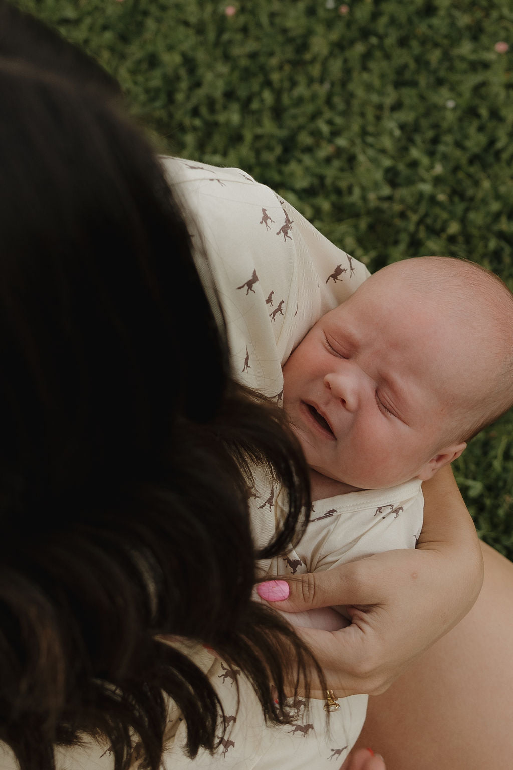 A dark-haired woman holds a newborn wrapped in a light, hypoallergenic blanket on grass. The baby, eyes closed and resting, wears forever french baby's Shortie Bamboo Zip | Wild Horses pajamas.