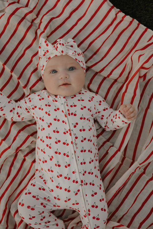 A baby with wide blue eyes lies on a red and white striped blanket, wearing a forever french baby outfit and the Bamboo Head Wrap | Sweetheart Cherries. Their arms are slightly raised as they look up.