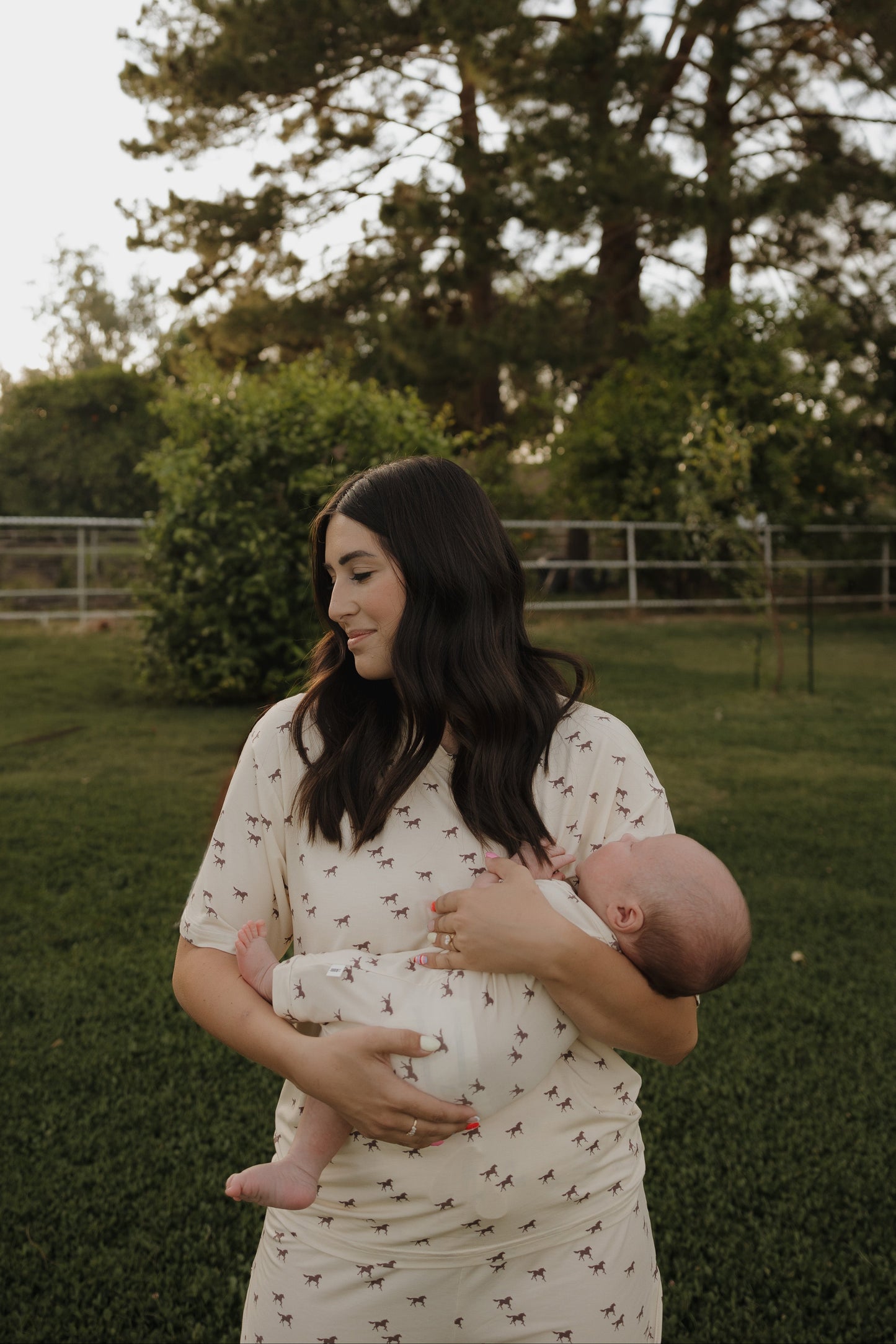 A woman with long dark hair stands on grass, gently cradling a baby dressed in the forever french baby Shortie Bamboo Zip | Wild Horses. She wears a light animal-print dress, with trees and greenery in the background.