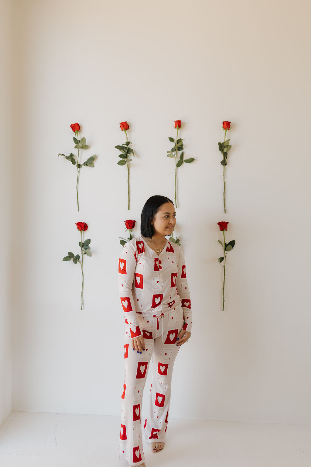 A woman stands against a white wall adorned with six vertical red roses, wearing the Love Day Women's Bamboo Pajamas by forever french baby, showcasing a stylish red heart pattern for ultimate comfort.