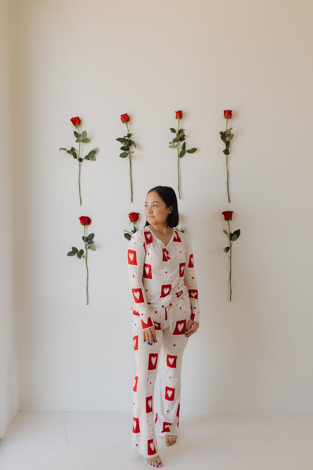 A person stands against a white wall decorated with red roses, wearing forever french baby's hypo-allergenic Women's Bamboo Pajamas | Love Day. The white outfit, adorned with red hearts and card patterns, adds elegance to the minimalist backdrop.