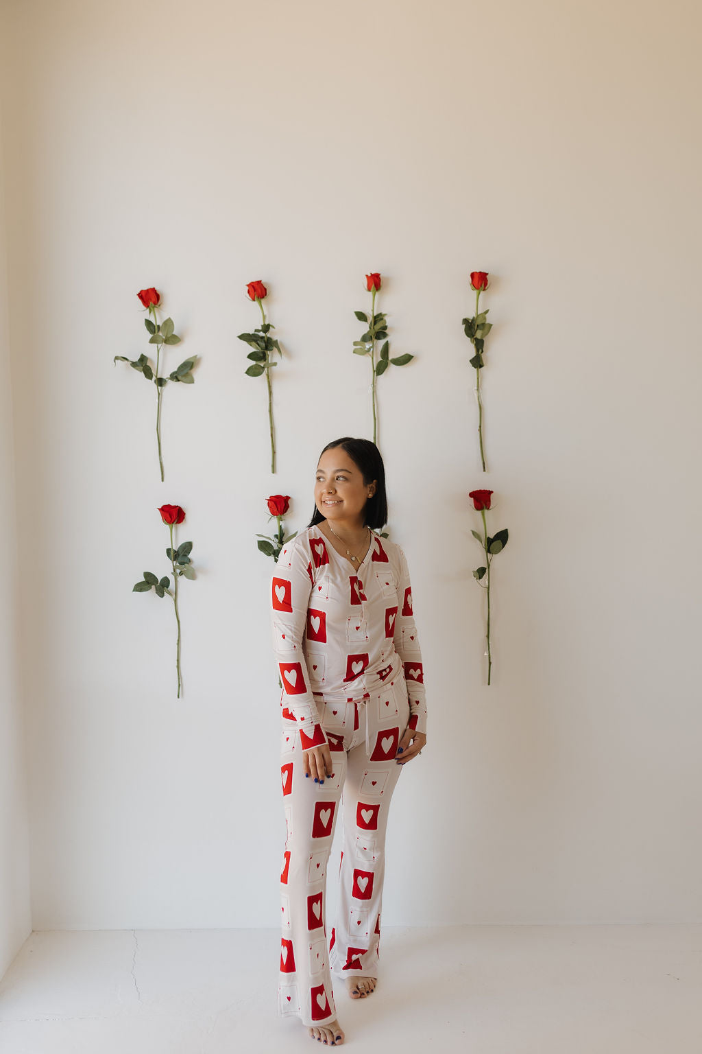 A person in forever french baby's Women's Bamboo Pajamas | Love Day, featuring red heart patterns, stands in a white room adorned with six red roses arranged in two vertical rows of three on the wall.