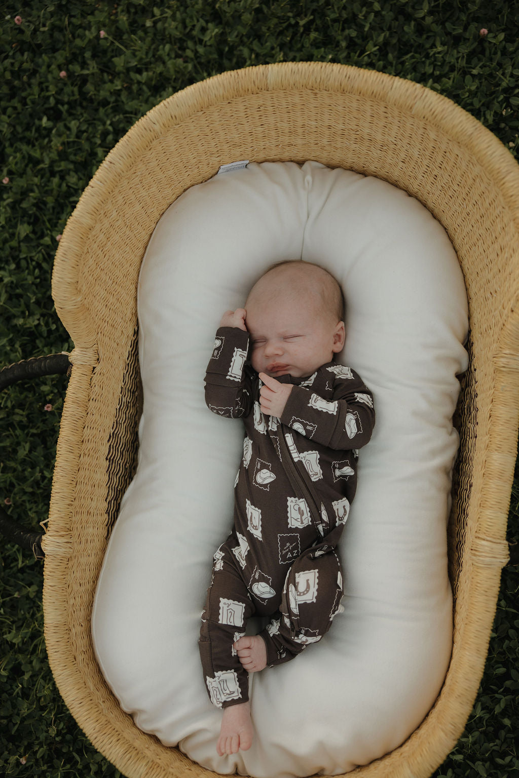 A newborn in forever french baby’s Bamboo Zip Pajamas | With Love from AZ sleeps soundly on a white cushion inside a woven basket, set on green grass.