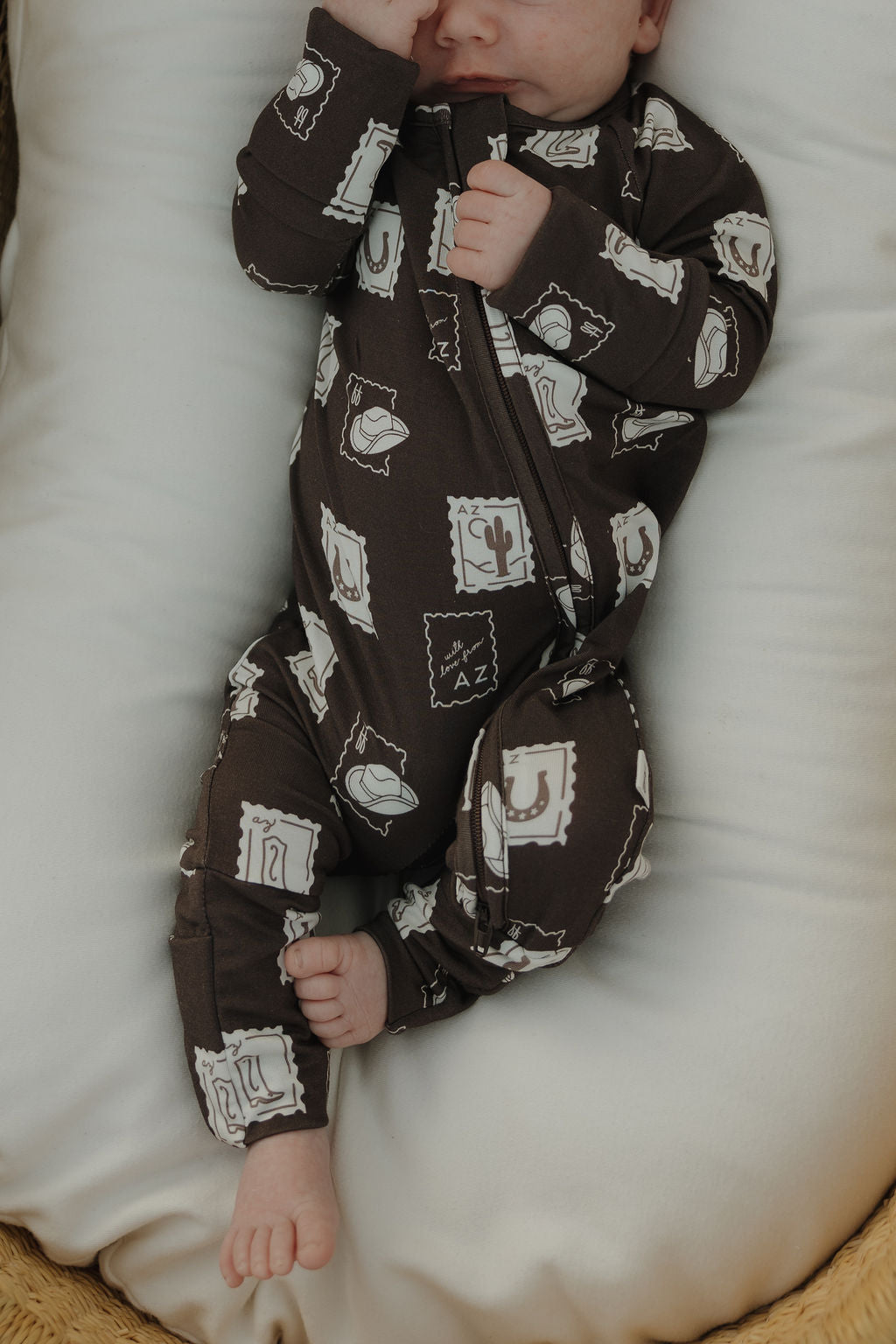 A baby in forever french baby's Bamboo Zip Pajamas | With Love from AZ—a brown zip-up onesie with white stamp and cactus prints—lies on a white pillow, one hand partly covering their face and bare feet showing.