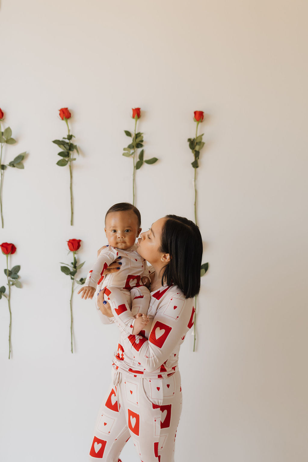 A woman gently kisses a baby while they both wear matching "Bamboo Zip Pajamas | Love Day" by forever french baby, featuring red heart designs on breathable fabric. A wall adorned with red roses stands behind them.