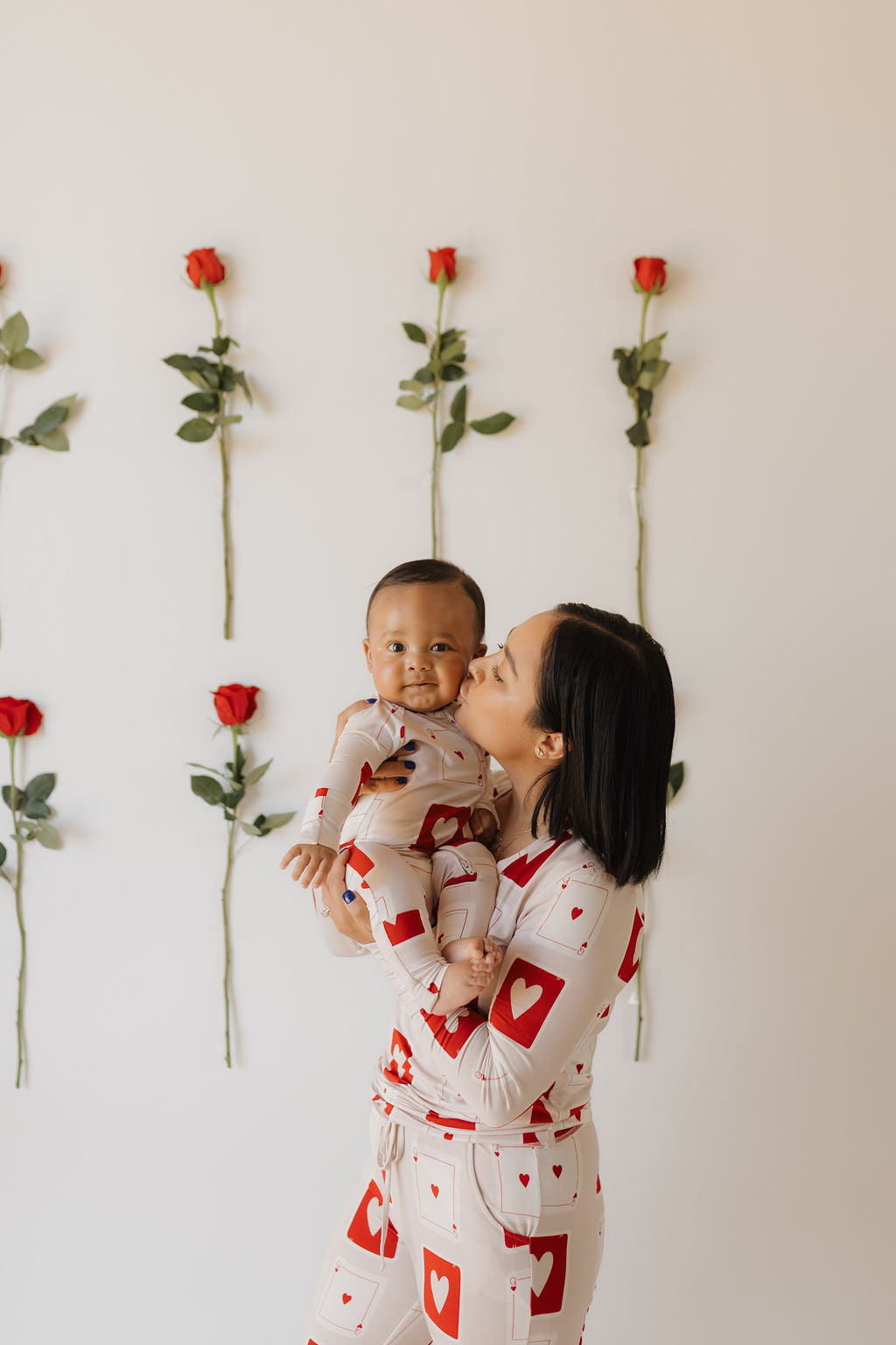 A woman embraces and kisses her smiling baby, both wearing forever french baby Bamboo Zip Pajamas in the Love Day design, featuring heart and square patterns made from breathable fabric. They stand before a white wall adorned with evenly spaced vertical red roses.