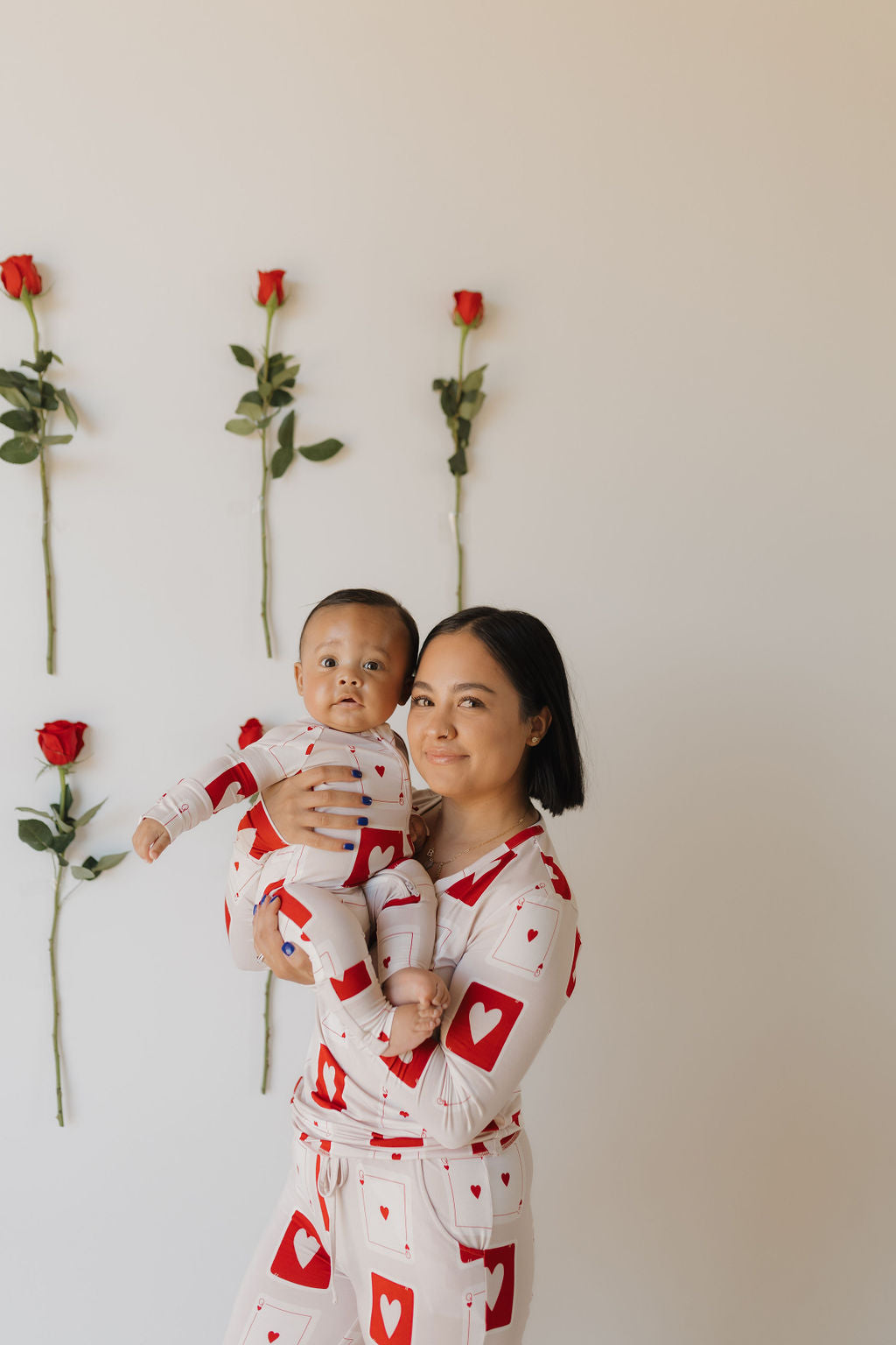 A woman and baby match in hypoallergenic "Bamboo Zip Pajamas | Love Day" by forever french baby, featuring red hearts. They pose against a light background with a wall lined with six vertical red roses.