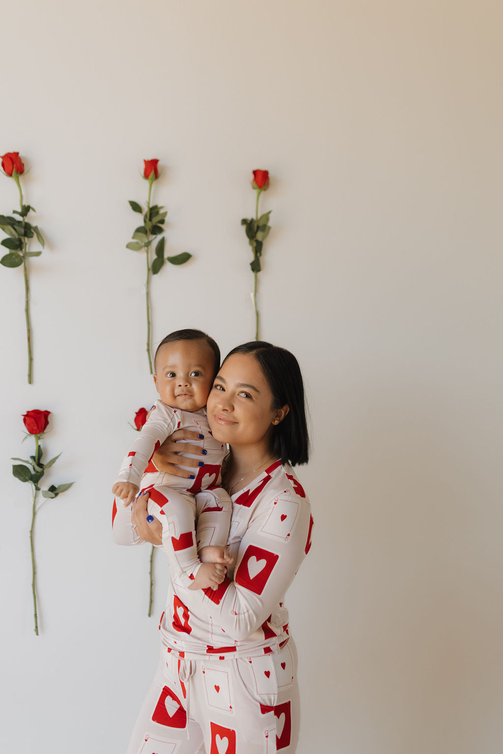 A woman in matching "Bamboo Zip Pajamas | Love Day" by forever french baby holds a similarly dressed, smiling baby. They pose joyfully against a beige wall adorned with five vertical red roses.