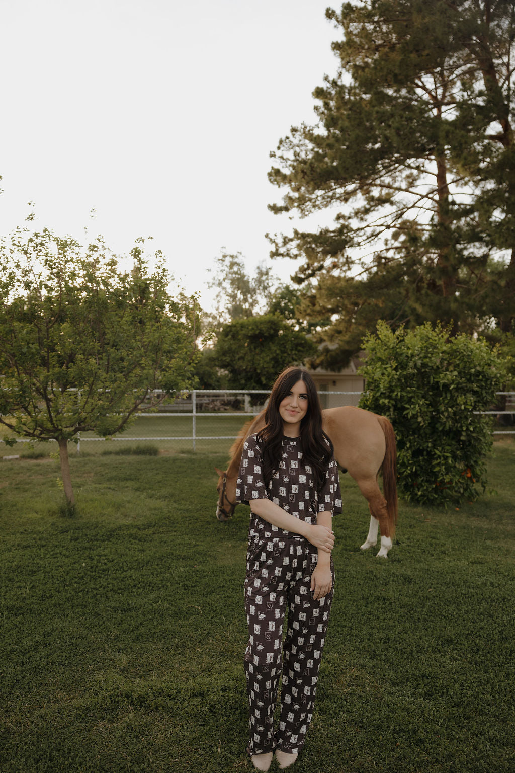 A woman with long dark hair wears the Women's Boxy Tee Lounge Set by forever french baby, standing on grass near trees. A horse grazes by a fence in the background, creating a calm, natural setting.