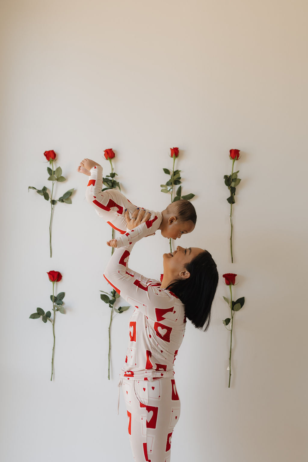 A woman wearing forever french baby's Bamboo Zip Pajamas | Love Day joyfully lifts a baby in matching attire against vertical red roses. The room exudes serenity and minimalist style, enhanced by the hypo-allergenic comfort of their clothing.