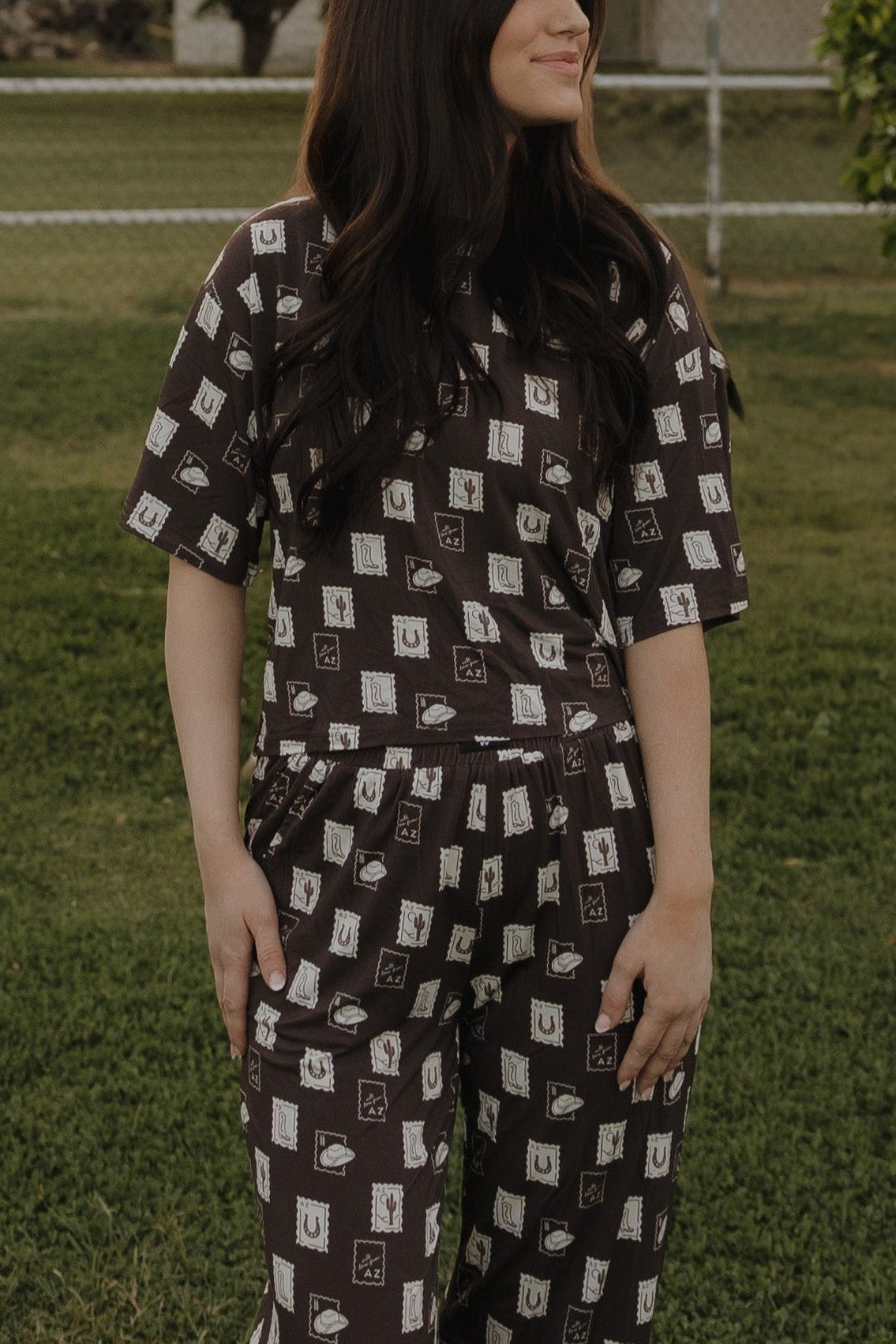 A person stands on grass wearing the forever french baby Women's Boxy Tee Lounge Set by With Love from AZ, featuring a black and white geometric pattern. Their long, dark hair is loose and their face is partially turned away; a fence appears in the background.
