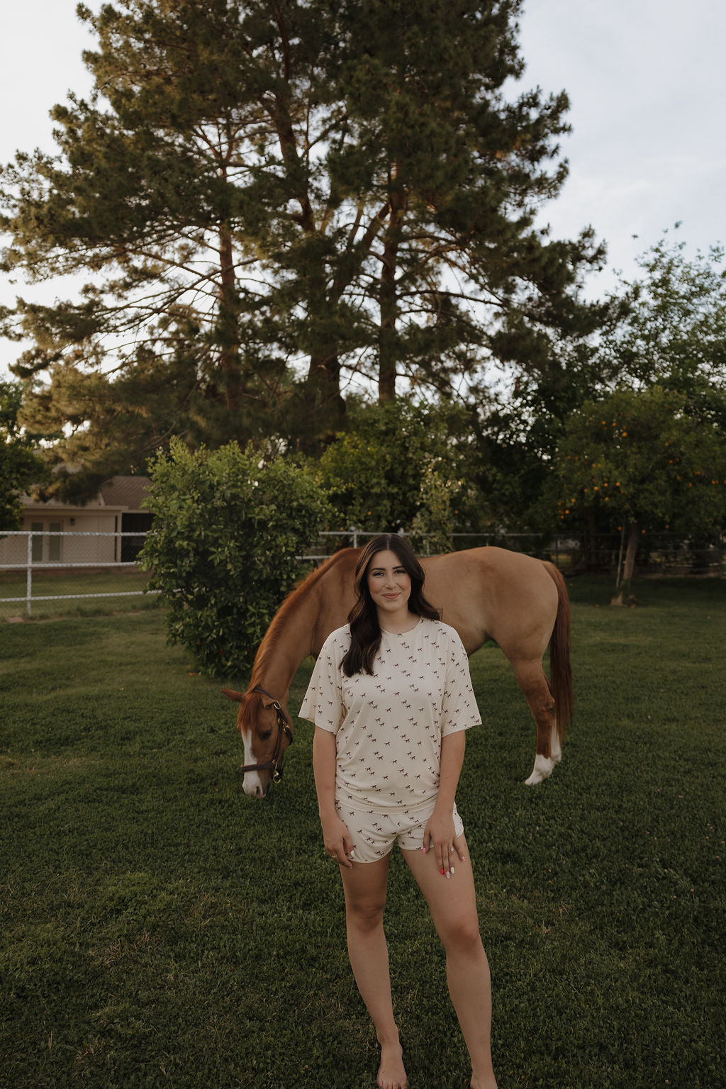A woman wearing the forever french baby Women's Bamboo Short Set | Wild Horses, light-colored and breathable, stands barefoot on grass smiling as a brown horse grazes nearby amid tall trees and a fenced yard, creating a tranquil scene.