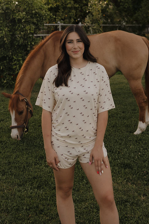 A woman with long dark hair stands on grass wearing the forever french baby Women's Bamboo Short Set | Wild Horses, featuring a breathable cream-colored design with small horse prints. In the background, a brown horse grazes among trees.