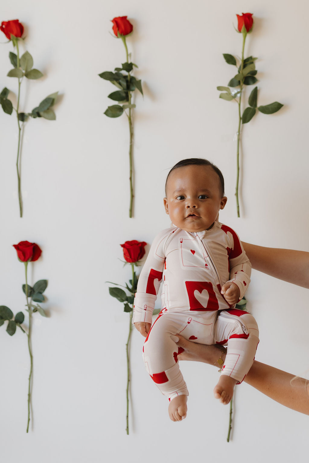 A baby is held up in front of a wall with red roses arranged vertically, wearing the "Bamboo Zip Pajamas | Love Day" by forever french baby. The red and white outfit, adorned with heart patterns, ensures breathable comfort and style.