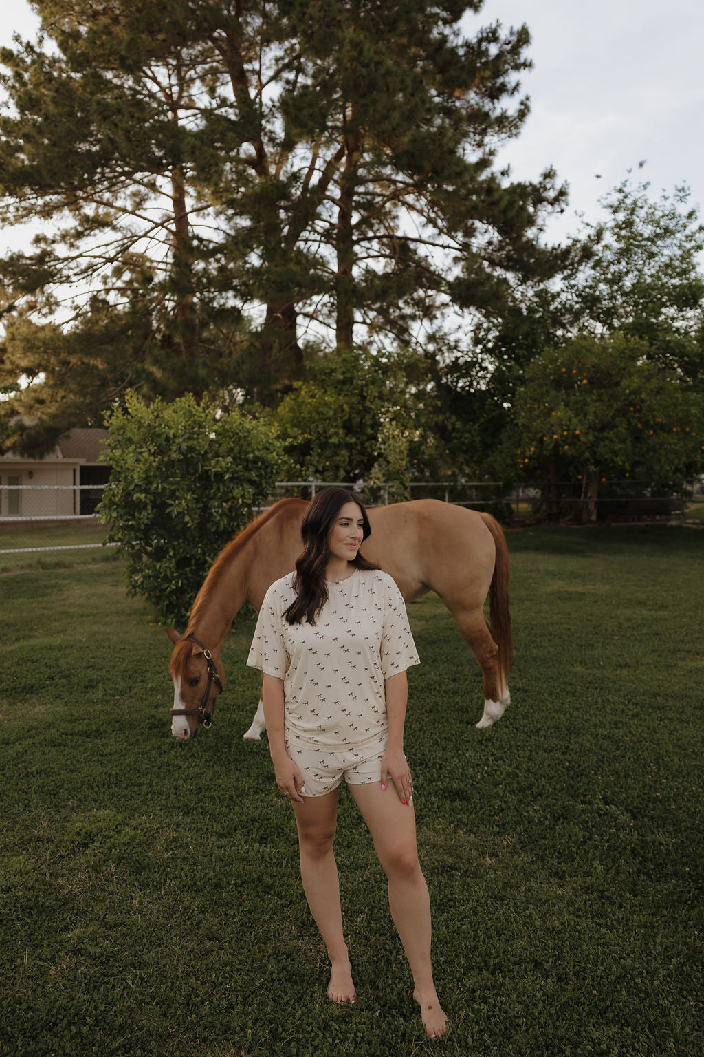 A woman in the forever french baby Women's Bamboo Short Set | Wild Horses stands barefoot on grass, smiling, with a brown horse grazing behind her. Tall trees and a house are visible in the background.
