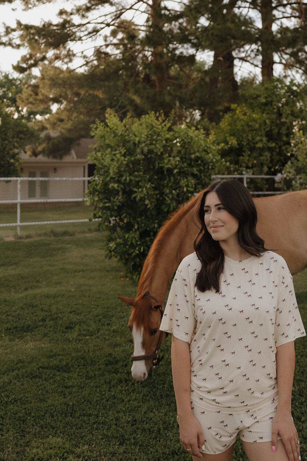 Wearing the forever french baby Women's Bamboo Short Set | Wild Horses, a woman with long dark hair smiles on the grass as a brown horse grazes nearby, enjoying the peaceful outdoors.