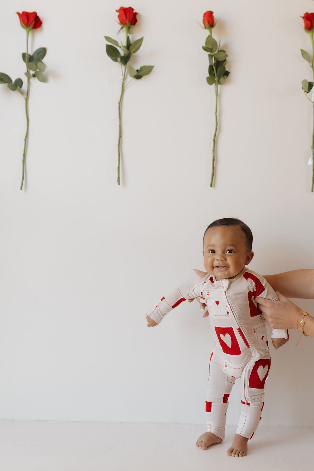 A smiling baby in forever french baby's Bamboo Zip Pajamas | Love Day is gently held up by someone’s hands. Four red roses on the white wall add charm to this hypo-allergenic clothing moment.