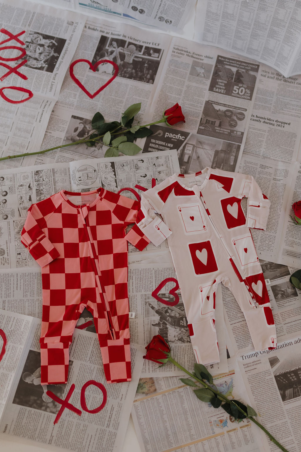 Two hypo-allergenic Bamboo Zip Pajamas with heart designs and red checkered patterns rest on newspaper sheets. Red roses, hand-drawn hearts, and "XO" enhance the romantic Valentine's theme, creating a charming scene perfect for showcasing breathable baby clothes by forever french baby.