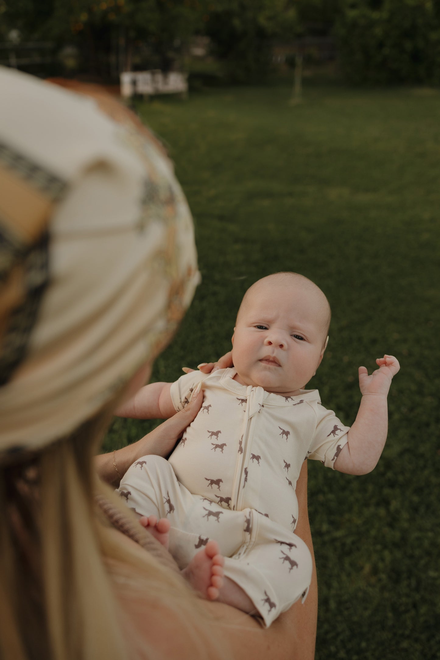 A baby wears the forever french baby Shortie Bamboo Zip in Wild Horses print while being held outdoors by an adult in a patterned headscarf, with grass and trees in the background.
