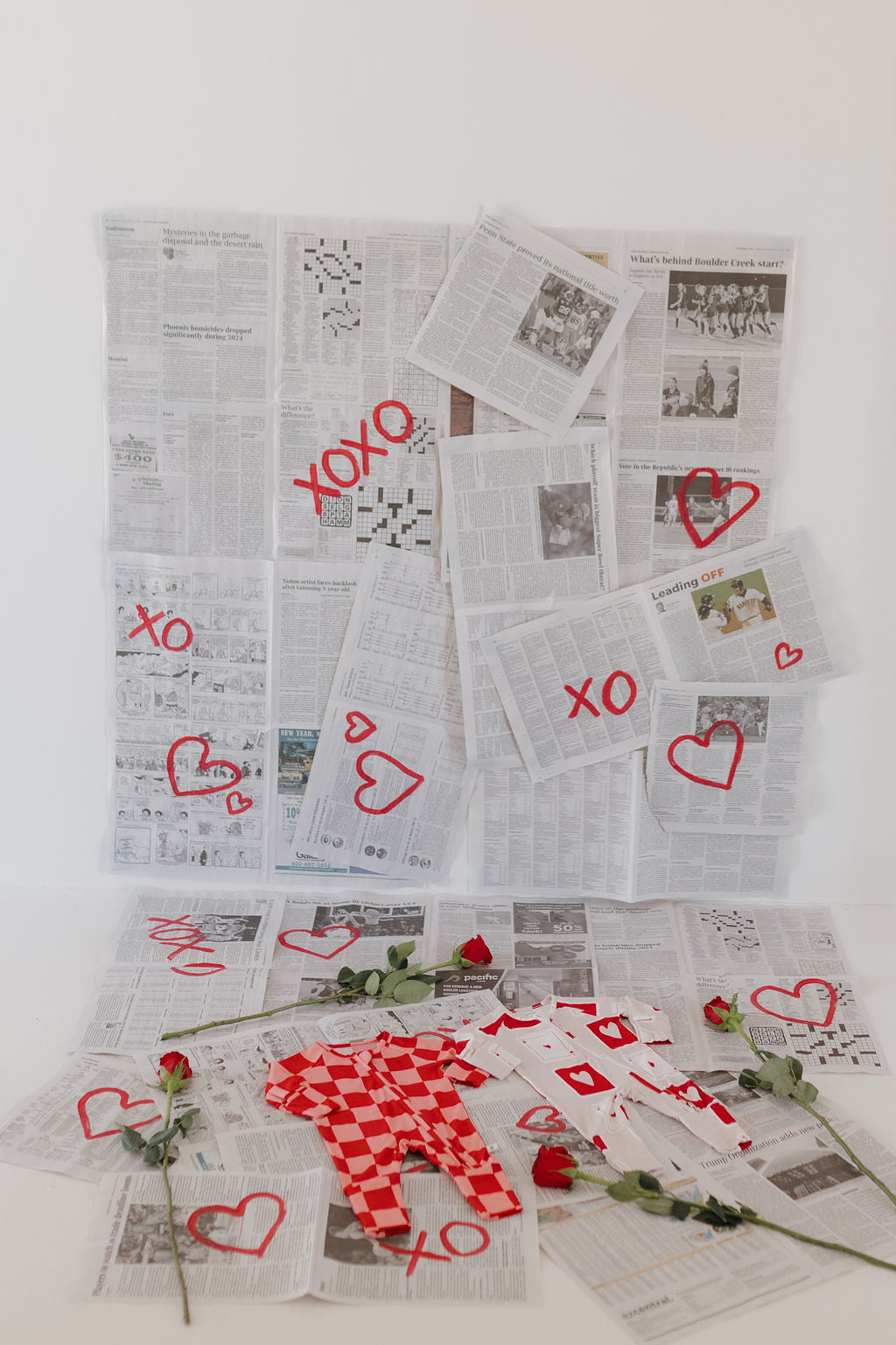 A wall adorned with newspapers, red hearts, and "xoxo" symbols creates a whimsical backdrop. Scattered newspapers, red roses, a red checkered garment, and hypo-allergenic comfort from forever french baby's Bamboo Zip Pajamas | XOXO complete the scene.