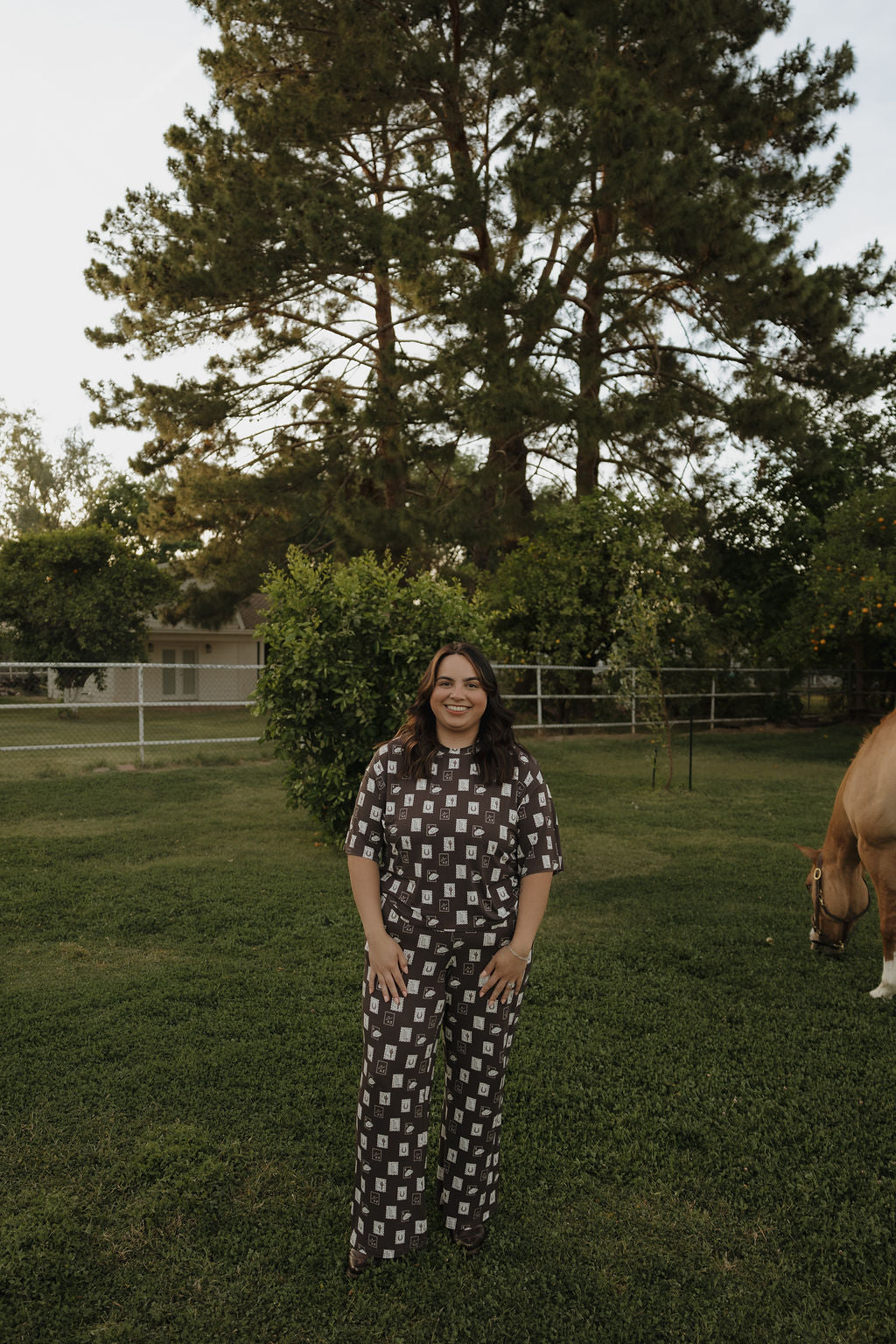 A woman smiles on green grass in front of a large tree, wearing the Women's Boxy Tee Lounge Set by forever french baby. A horse grazes nearby, with a house and fence visible in the background.