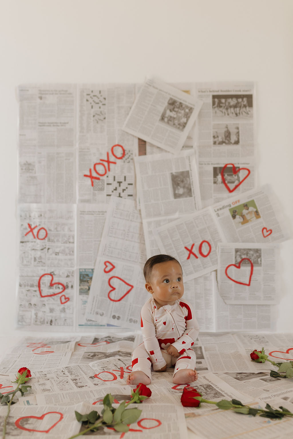 A baby sits on a floor covered with newspaper pages, wearing the forever french baby Bamboo Zip Pajamas | Love Day, featuring red hearts. The background wall is also covered in newspapers with "xoxo" and red hearts drawn on them. Red roses are scattered across the floor.