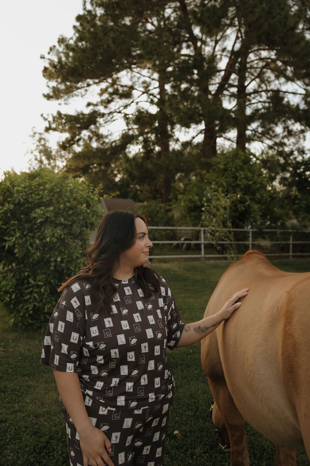 A woman wearing the Women's Boxy Tee Lounge Set by forever french baby stands on grass, gently touching a brown cow, surrounded by trees and lush greenery.