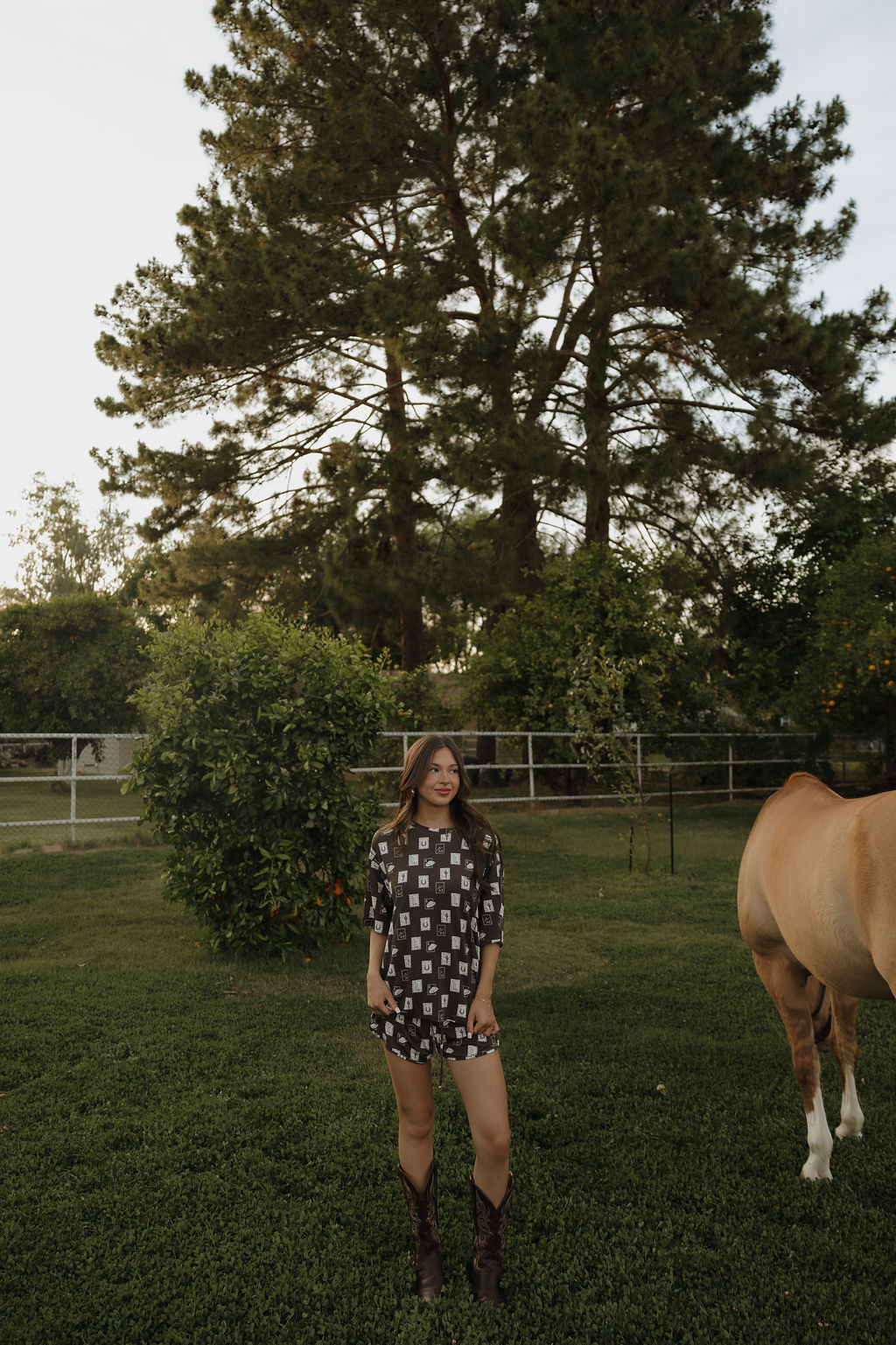 A woman in a forever french baby Women's Bamboo Short Set stands on grass near a brown horse, with trees and a fence in the background on a sunny day.
