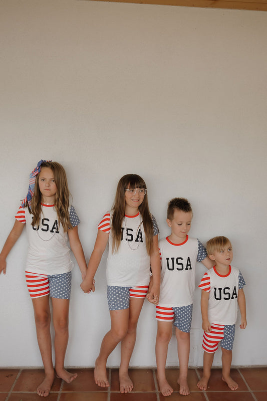 Four barefoot children stand against a white wall, wearing forever french baby’s Bamboo Short Set | Stars & Stripes. The matching outfits feature red, white, and blue stripes with star patterns. Two kids are holding hands.