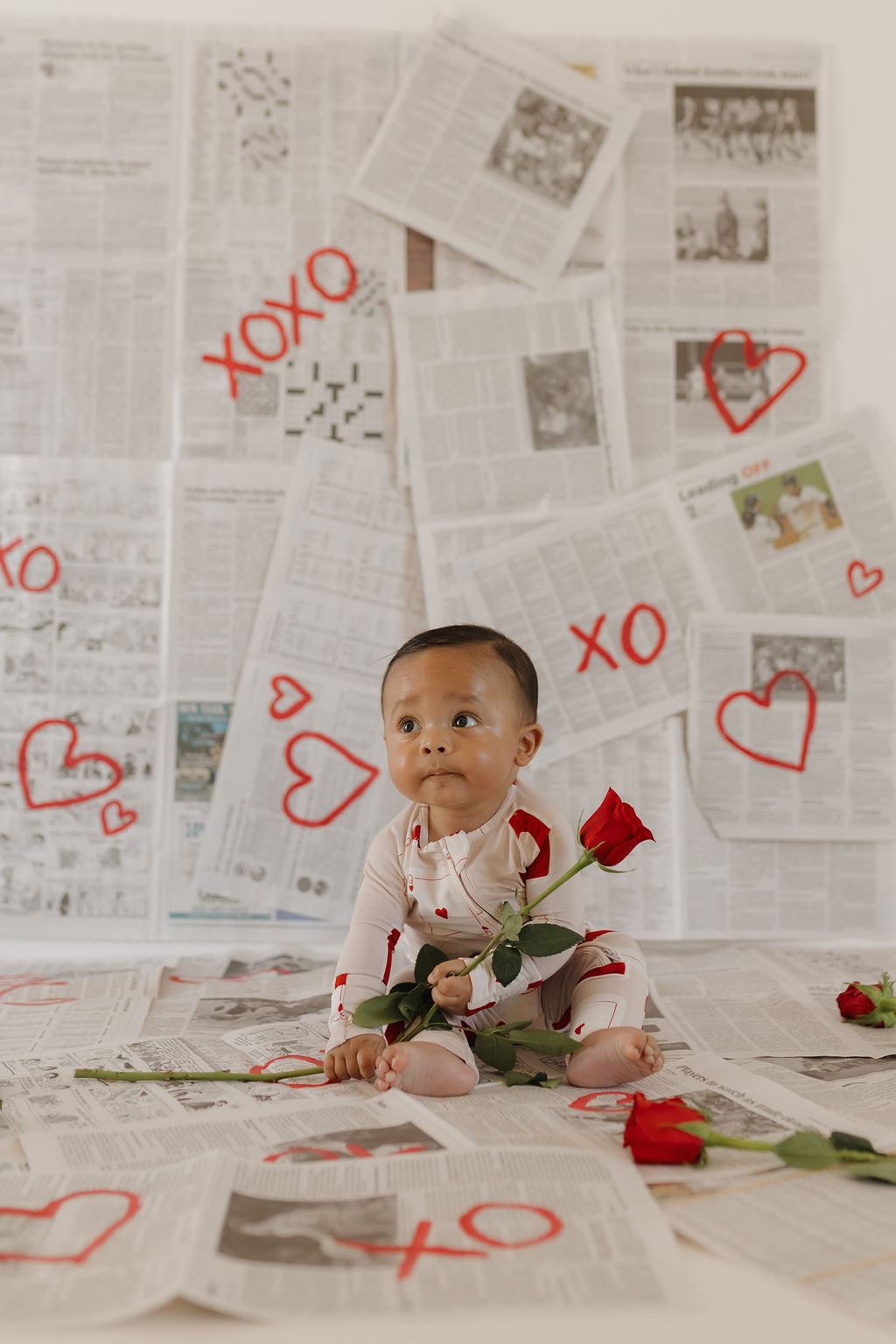 A baby in forever french baby's Bamboo Zip Pajamas | Love Day, featuring heart patterns, sits safely on a newspaper-covered floor with scattered roses and a red rose in hand. The background displays newspapers adorned with red heart and "XOXO" drawings.